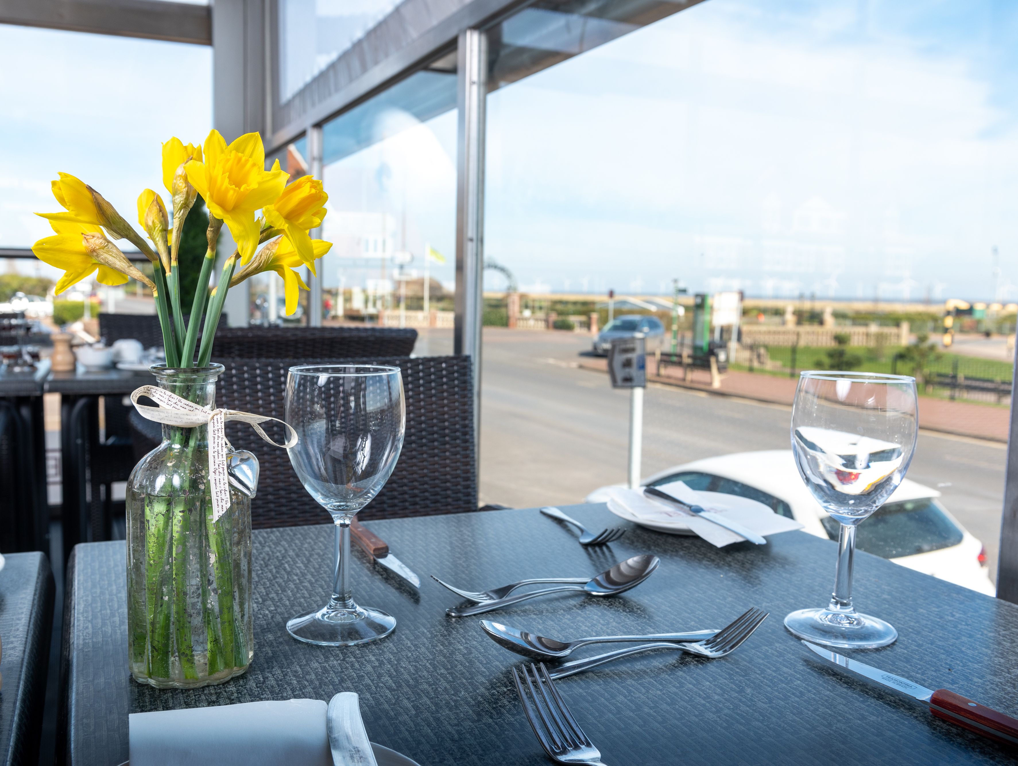 Table set for two with daffodils in a glass vase, overlooking a bright street through large windows.
