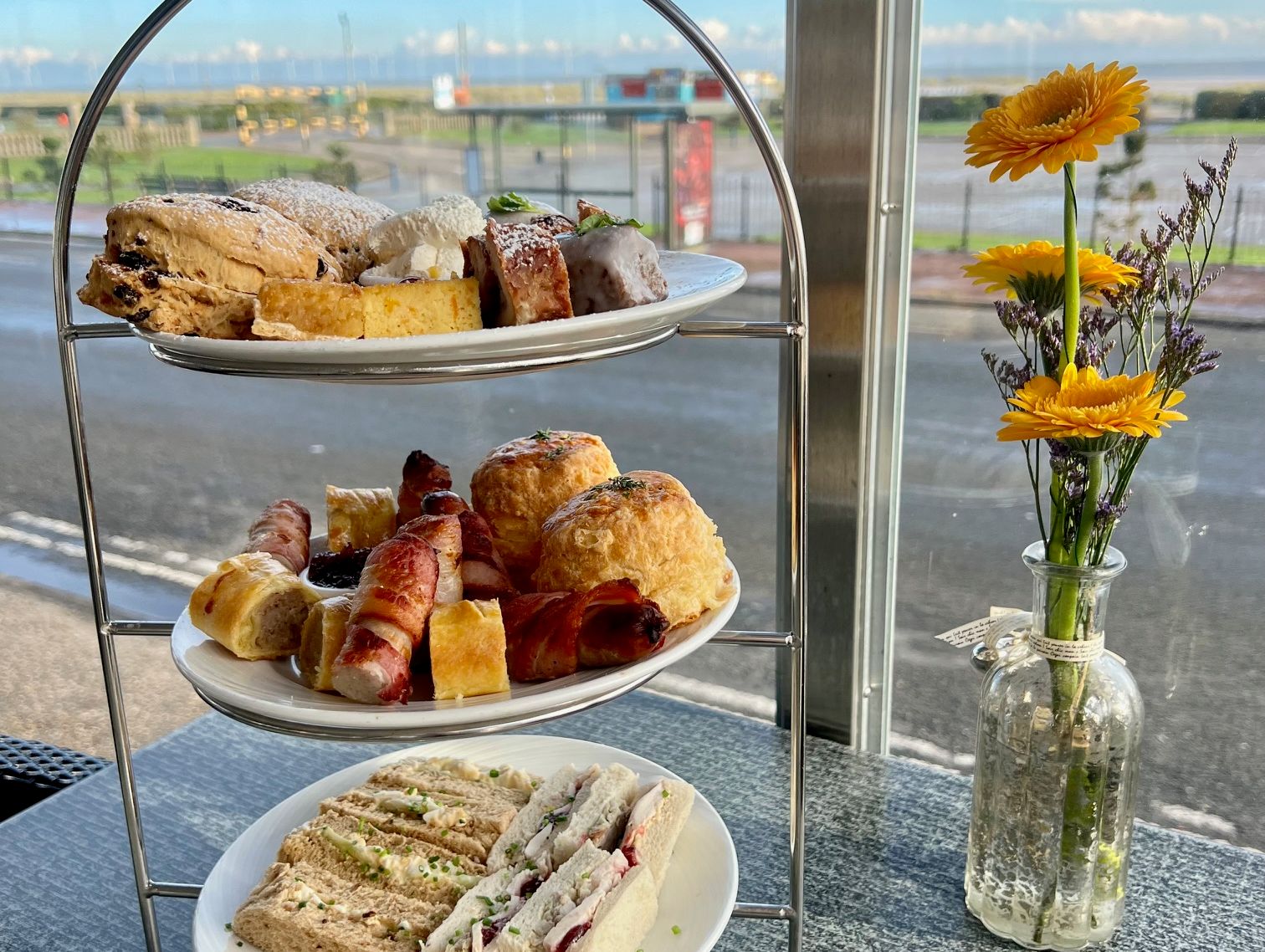 Three-tier afternoon tea stand with assorted sandwiches, scones, pastries, and cakes next to a vase of yellow flowers by a window.