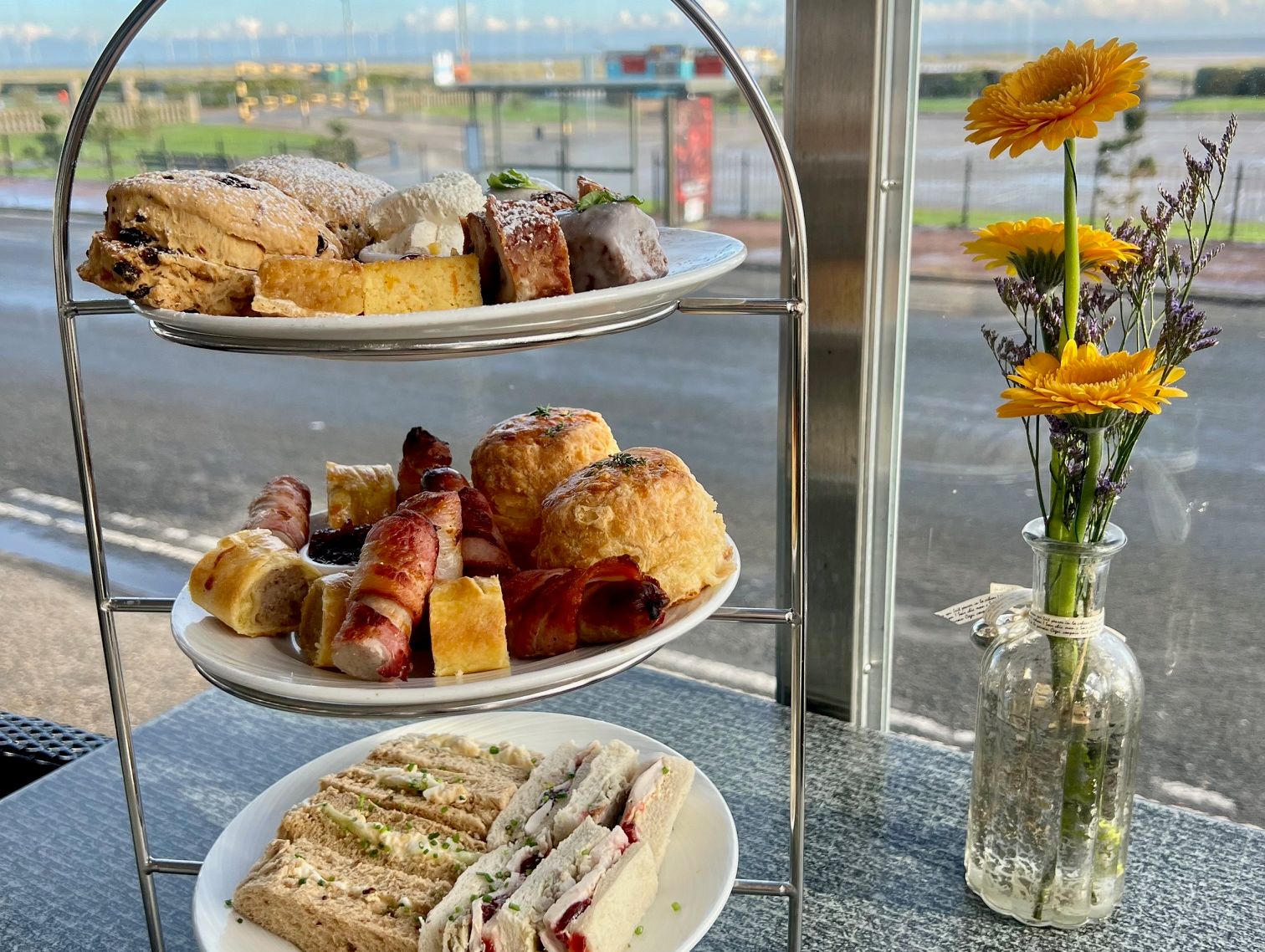 Three-tier afternoon tea stand with assorted sandwiches, scones, pastries, and cakes next to a vase of yellow flowers by a window.