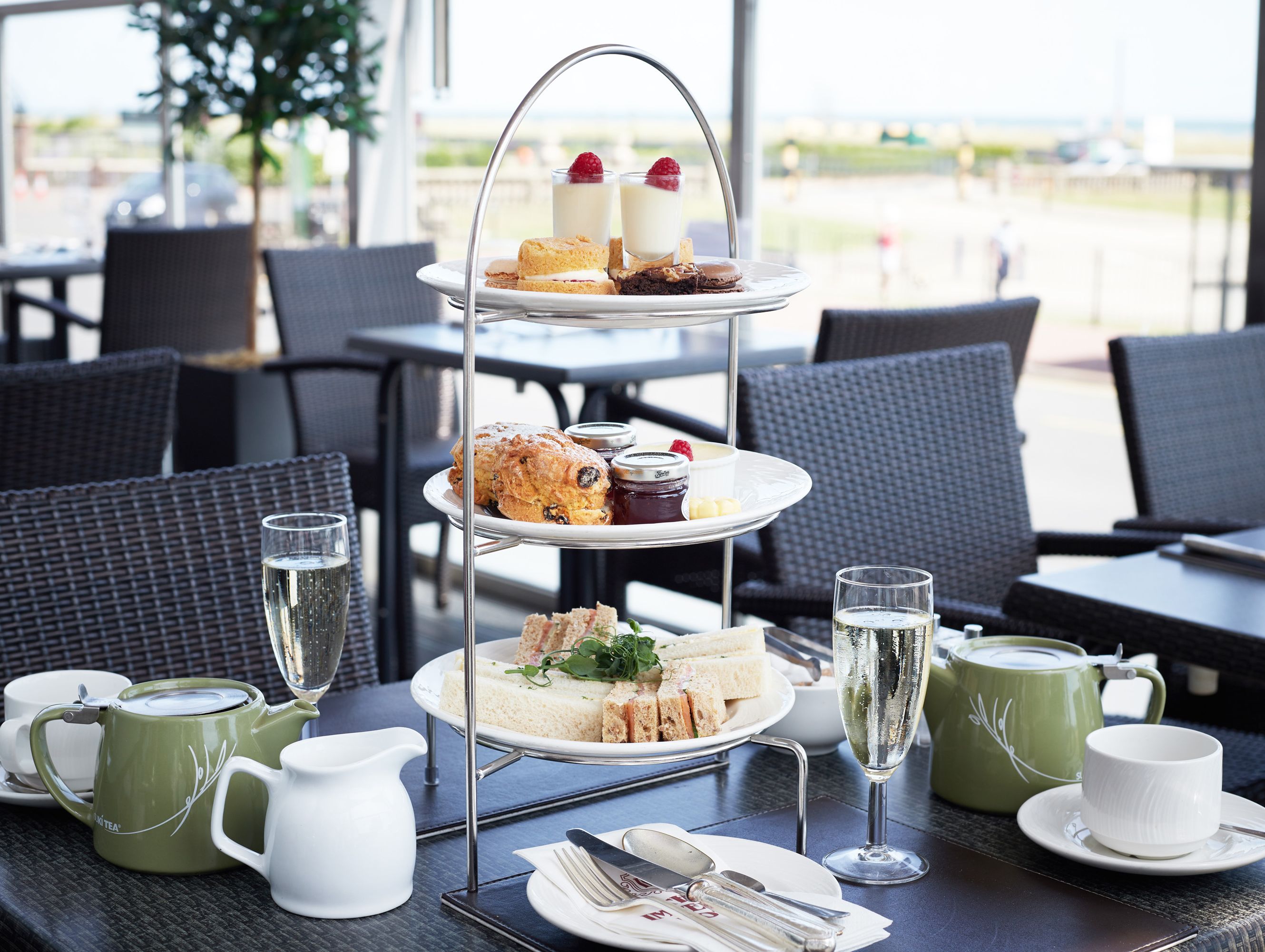 Three-tier afternoon tea set with sandwiches, scones, and desserts, accompanied by tea pots and glasses of champagne on an outdoor restaurant table.