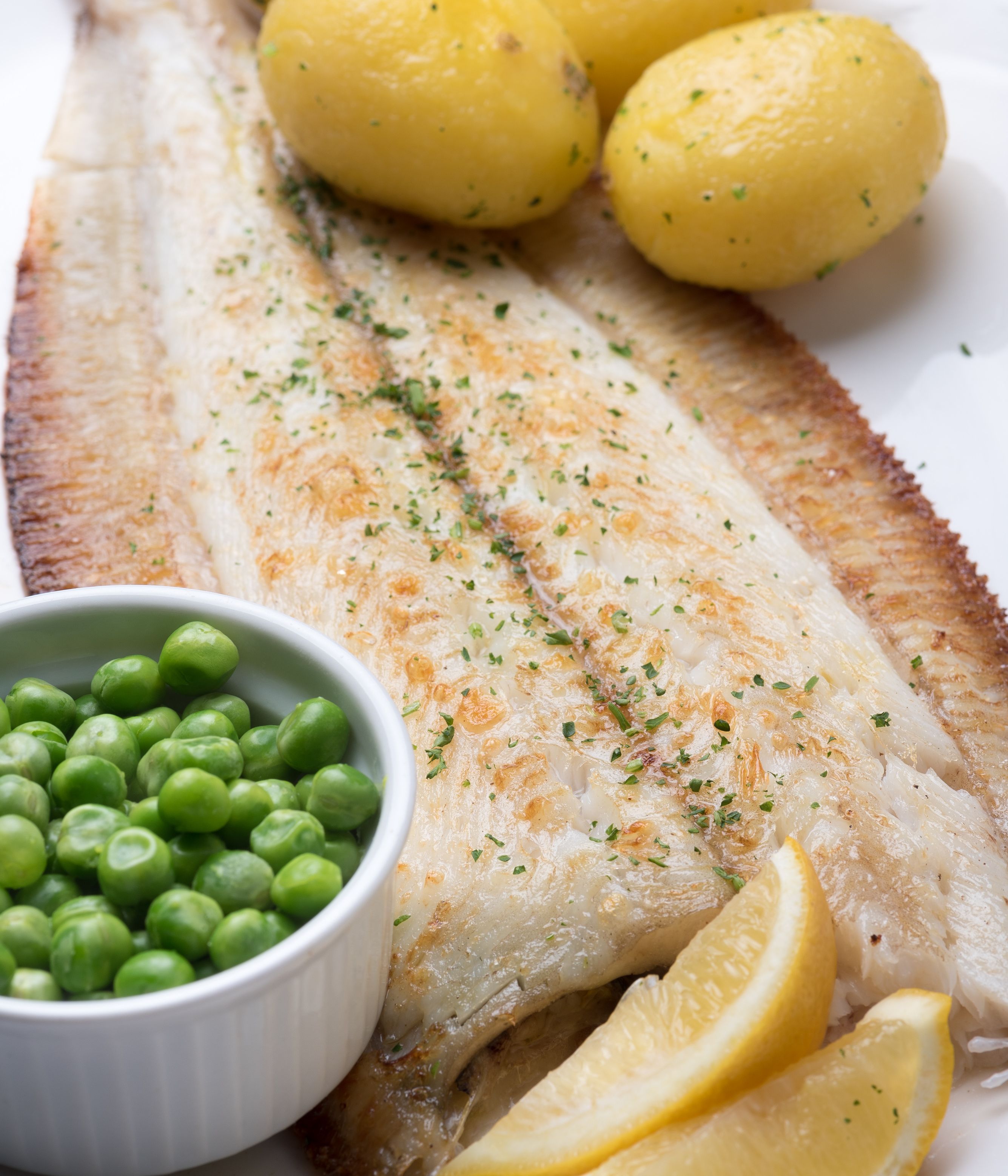 Grilled fish fillet served with boiled potatoes, green peas, and lemon wedges on a white plate