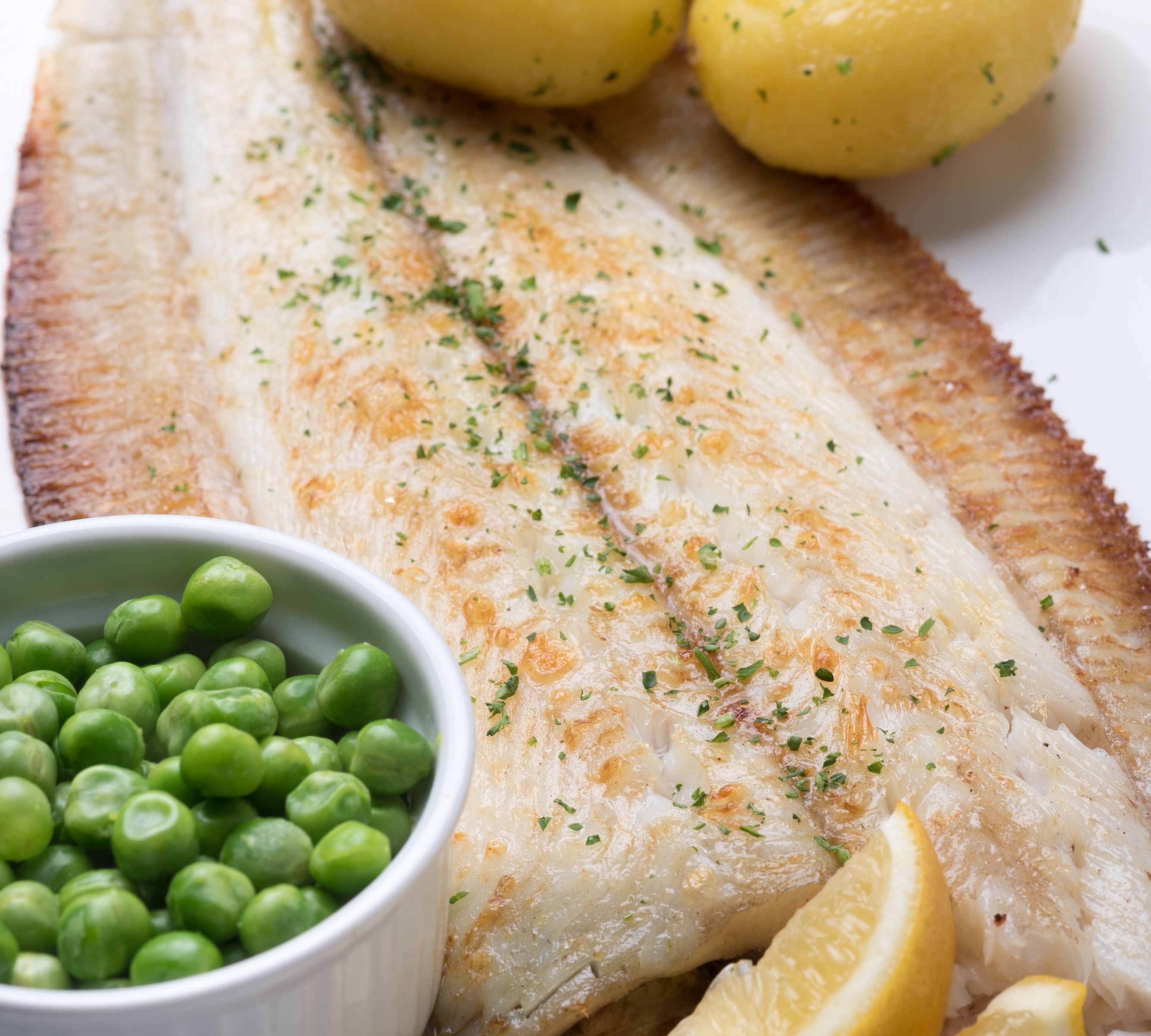Grilled fish fillet served with boiled potatoes, green peas, and lemon wedges on a white plate