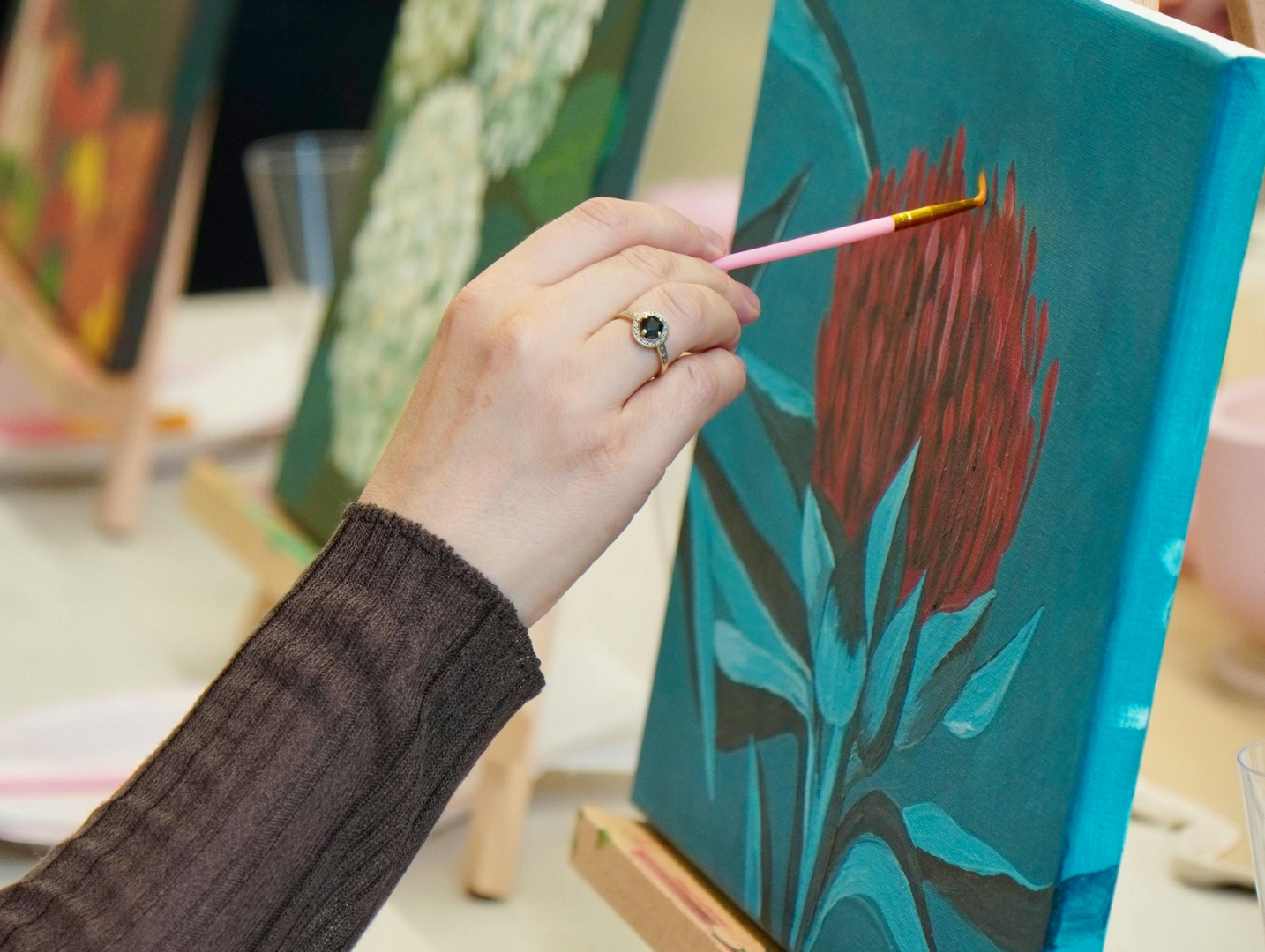 Close-up of a person painting a red flower on a canvas on an easel during a workshop