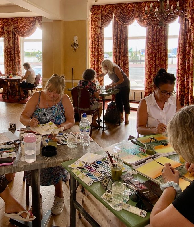 People painting with watercolours at tables in a sunlit room with patterned curtains and chandeliers