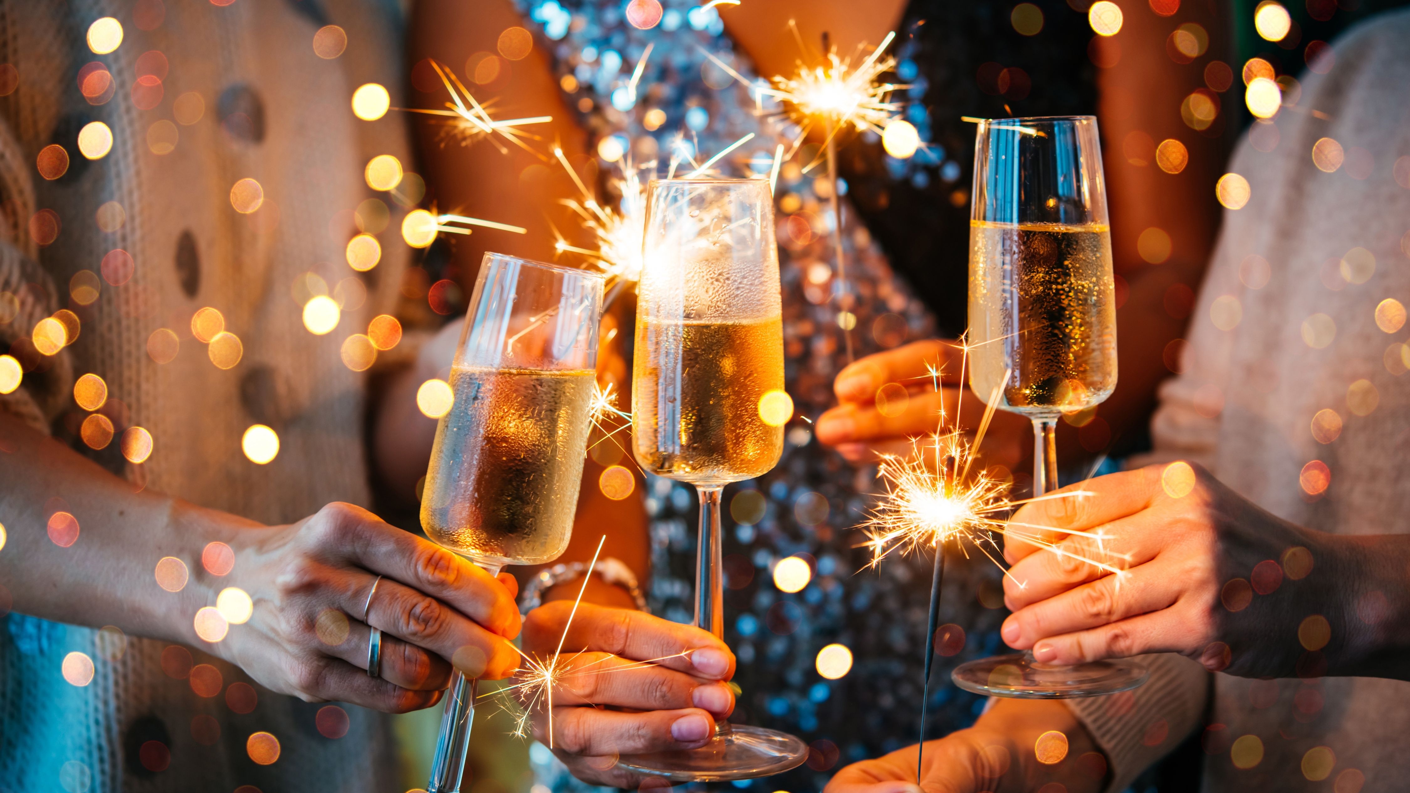 Hands holding champagne flutes and lit sparklers at a celebration with colourful bokeh lights