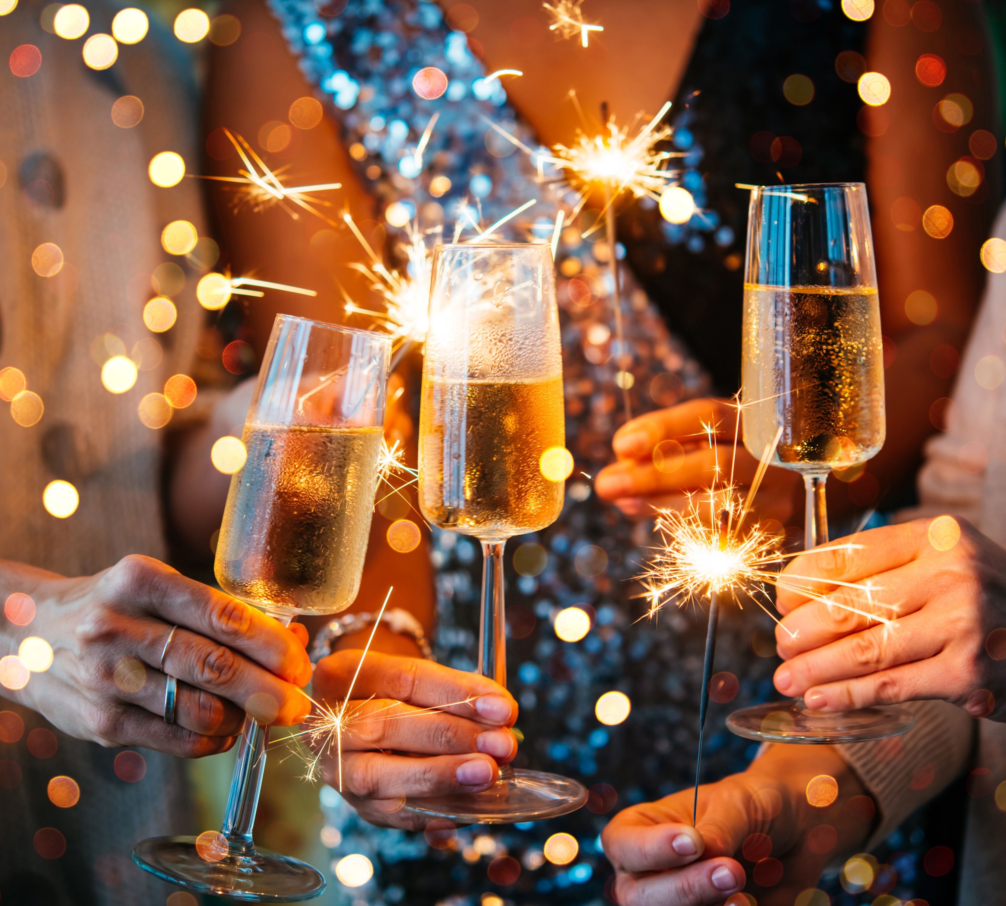 Hands holding champagne flutes and lit sparklers at a celebration with colourful bokeh lights