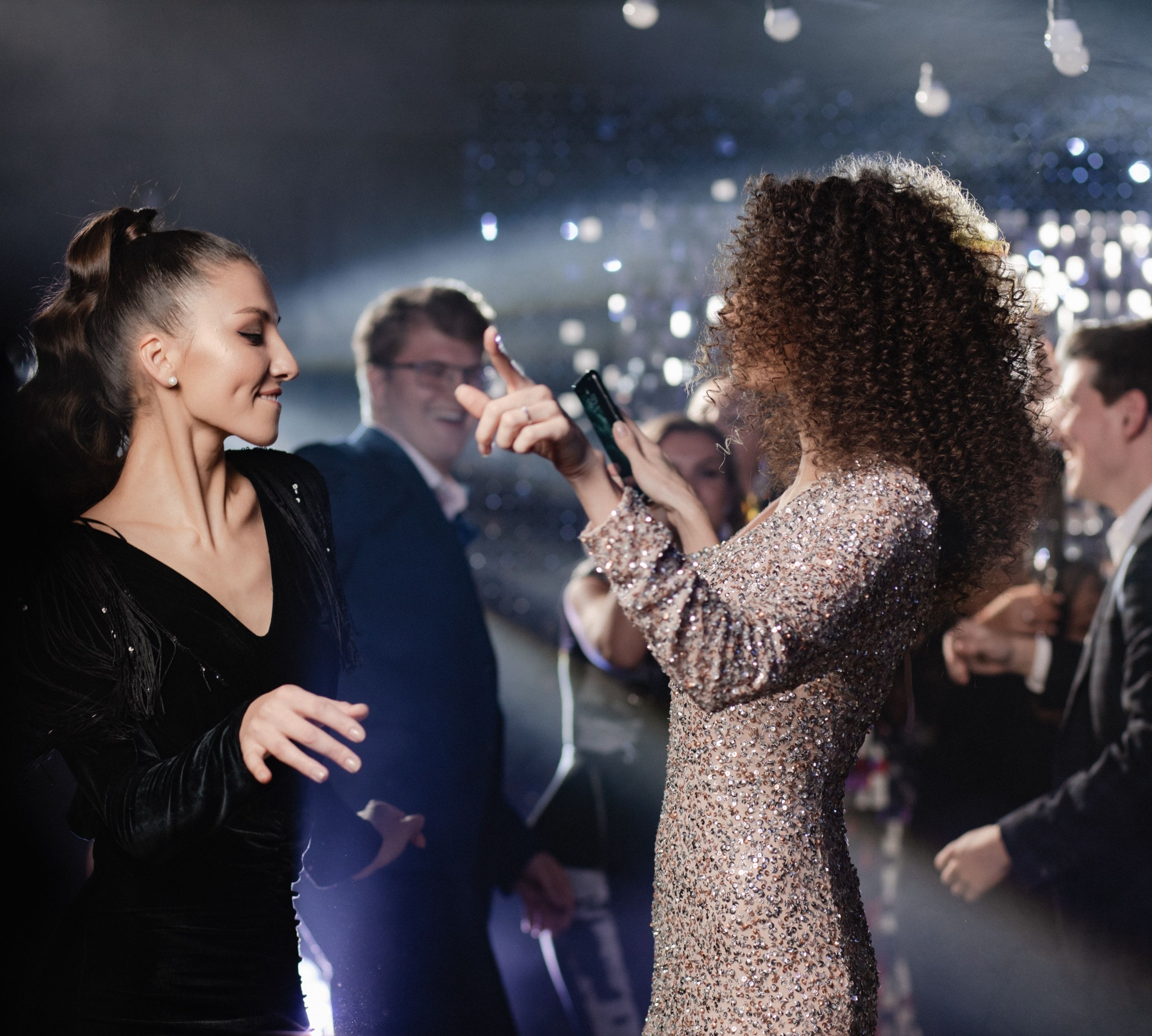Two women dancing at a party with sparkling lights and guests in the background