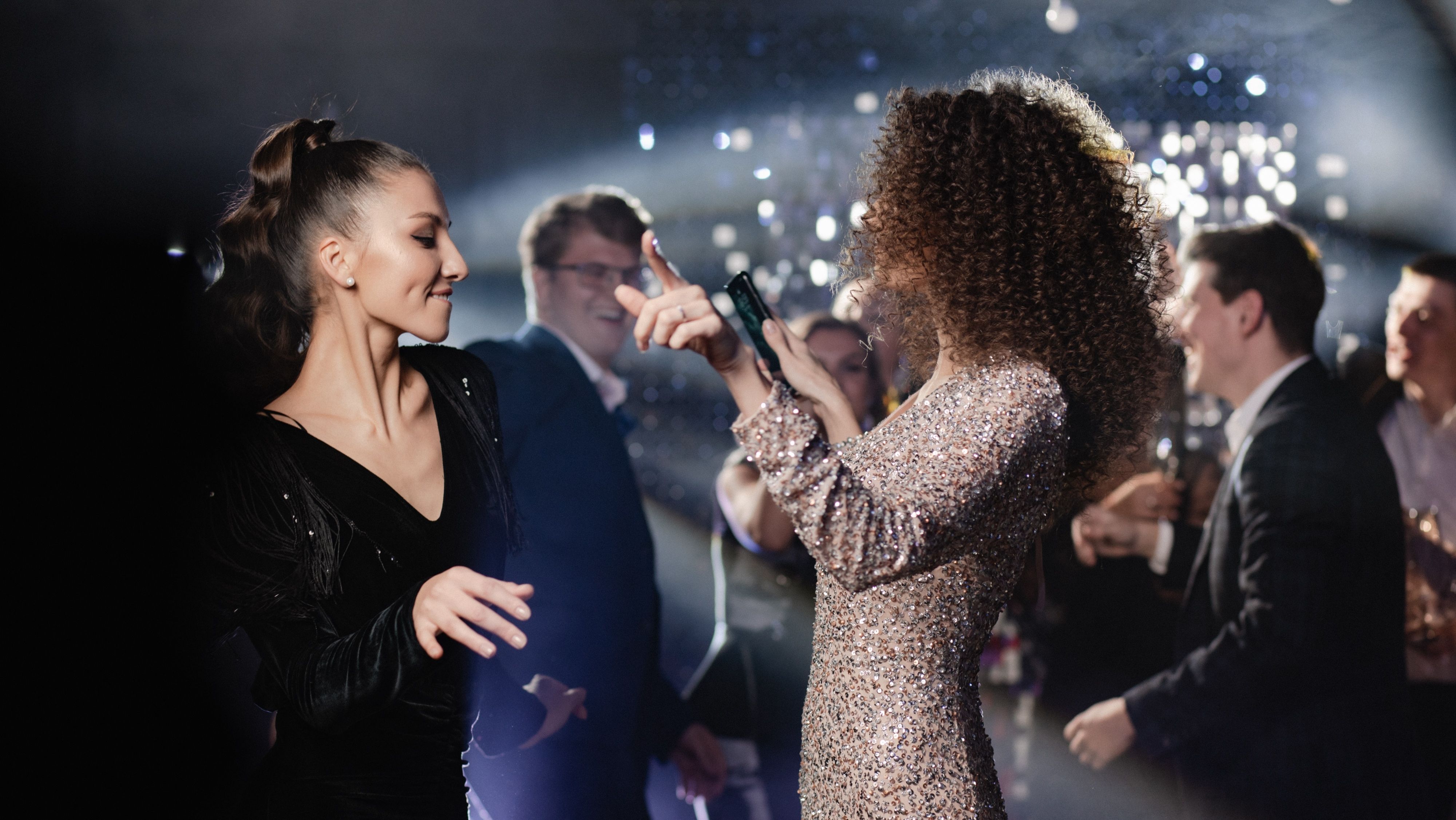 Two women dancing at a party with sparkling lights and guests in the background
