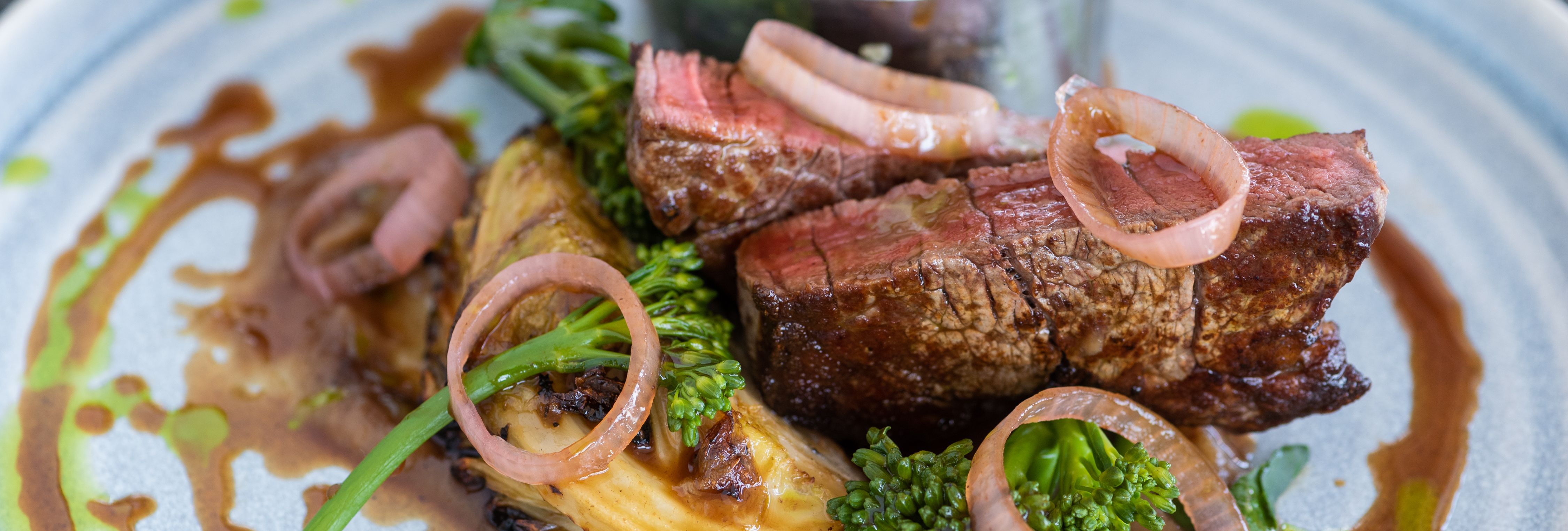 Plated gourmet meal with sliced steak, broccolini, roast potatoes, pickled onions, and sauce.