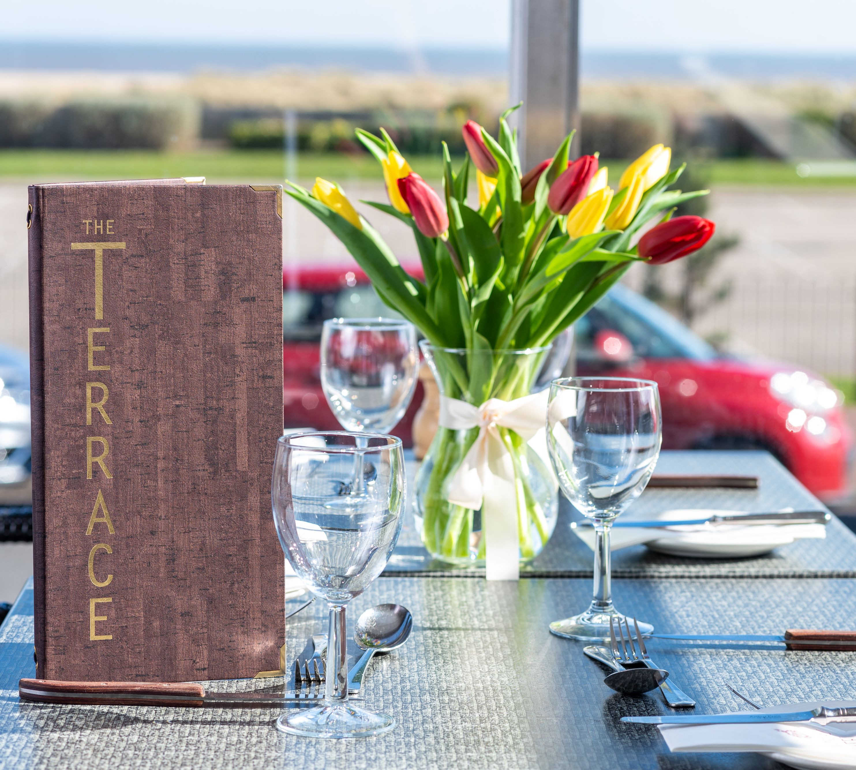 Outdoor restaurant table set with glassware, cutlery, a menu labeled 'The Terrace', and a vase of red and yellow tulips.