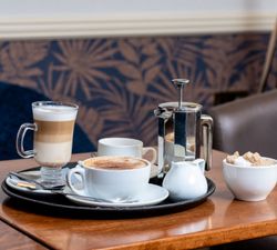 A tray with coffee, a latte, a French press, a jug of milk, and a bowl of sugar cubes on a wooden table.