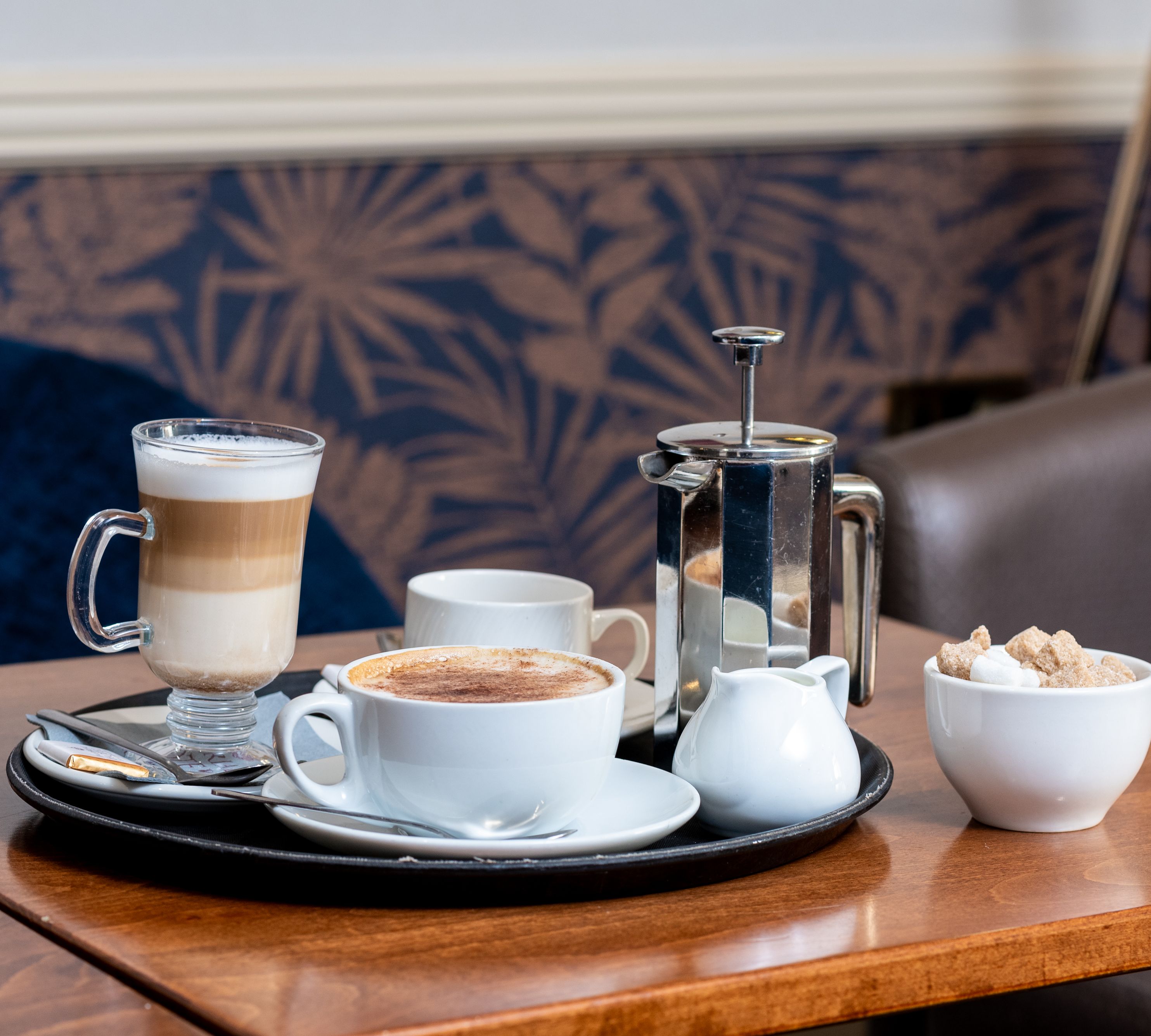 A tray with coffee, a latte, a French press, a jug of milk, and a bowl of sugar cubes on a wooden table.
