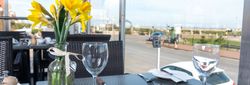 Close-up view of a table set for dining with yellow flowers in a vase and glassware, with a road and seaside view through large windows.