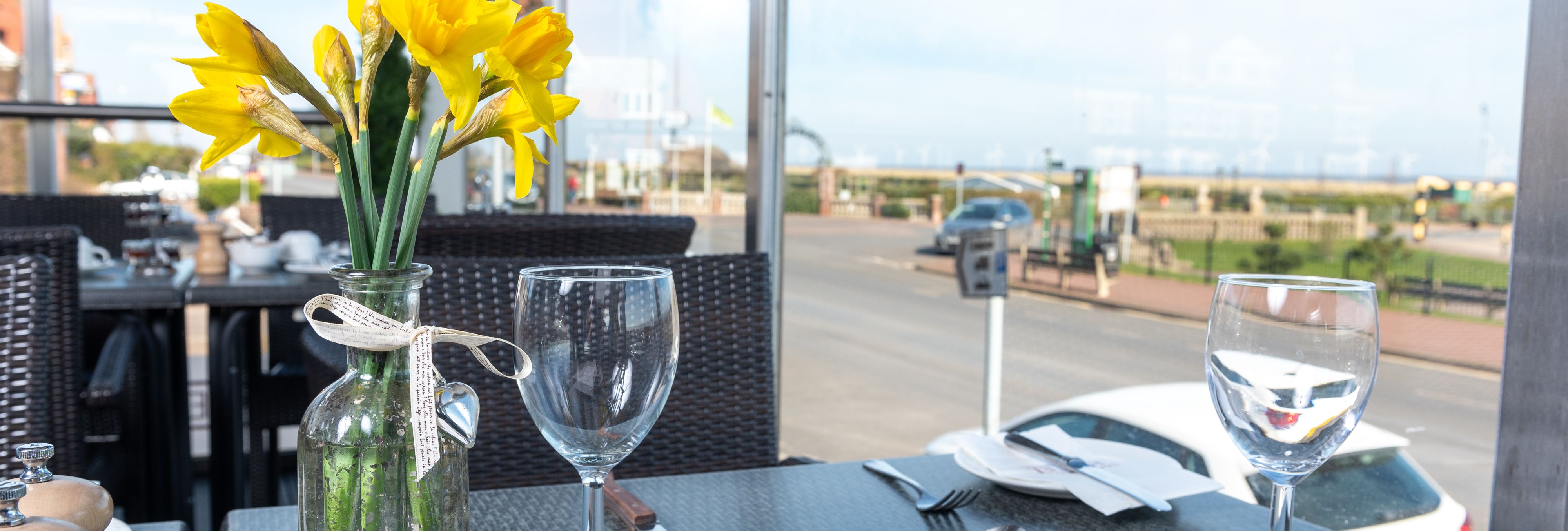 Close-up view of a table set for dining with yellow flowers in a vase and glassware, with a road and seaside view through large windows.