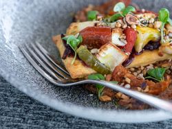 Close-up of a gourmet vegetable tart with roasted vegetables, cheese, microgreens, and seeds on a textured plate with a fork.