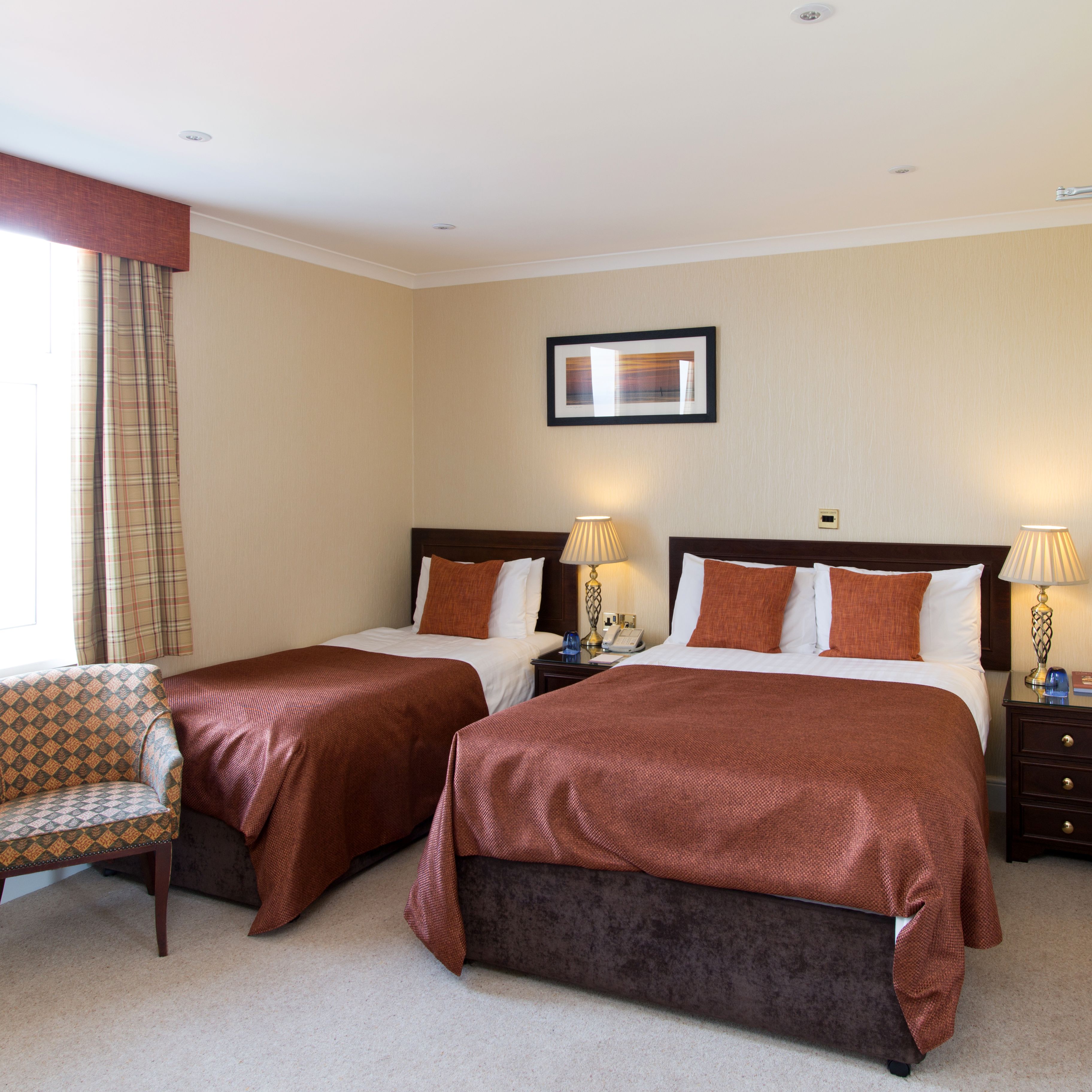 Bright hotel bedroom with double and single beds, bedside lamps, plaid curtains and upholstered chair