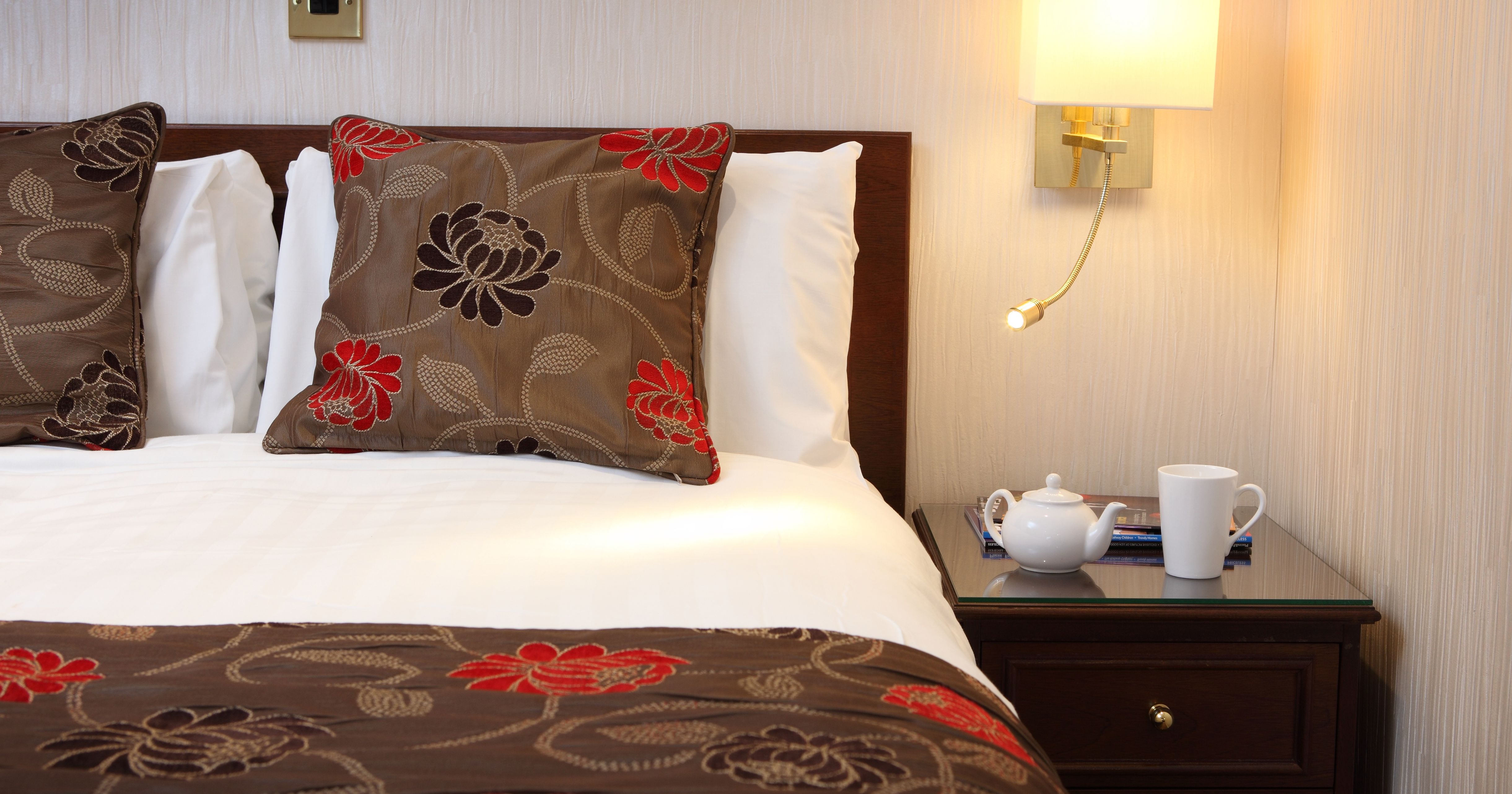 Hotel bed with floral cushions, bedside lamp, and teapot on a nightstand
