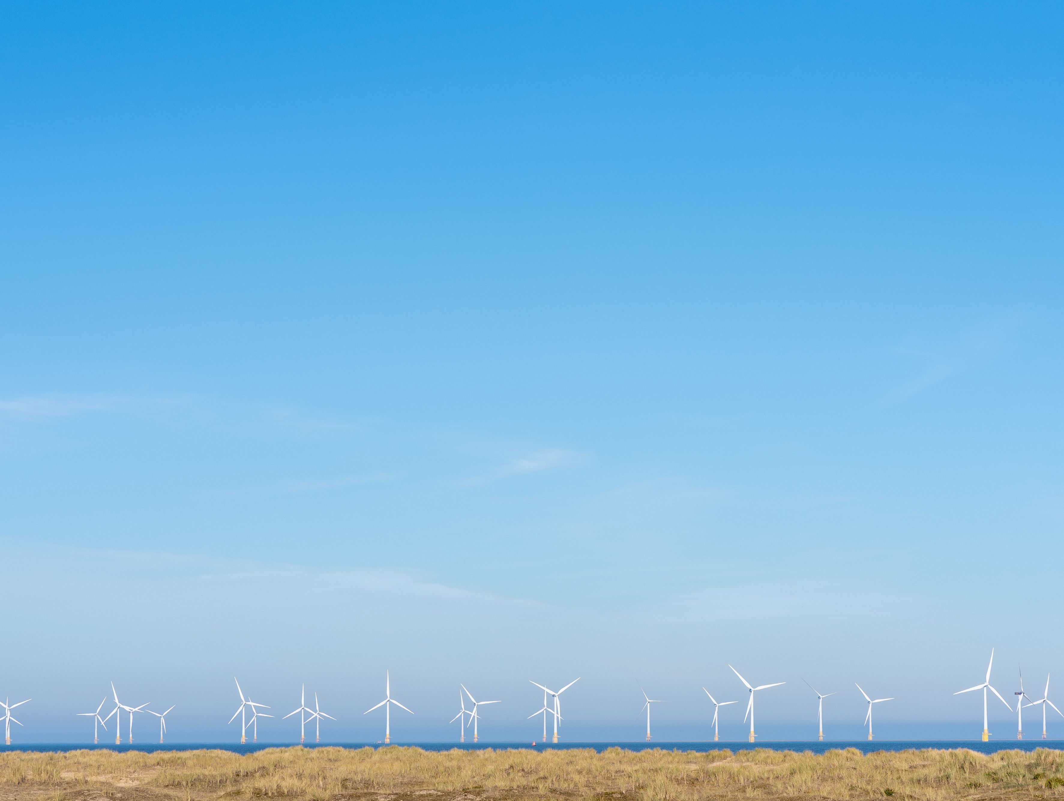Offshore wind turbines on the horizon beyond sandy dunes under a clear blue sky