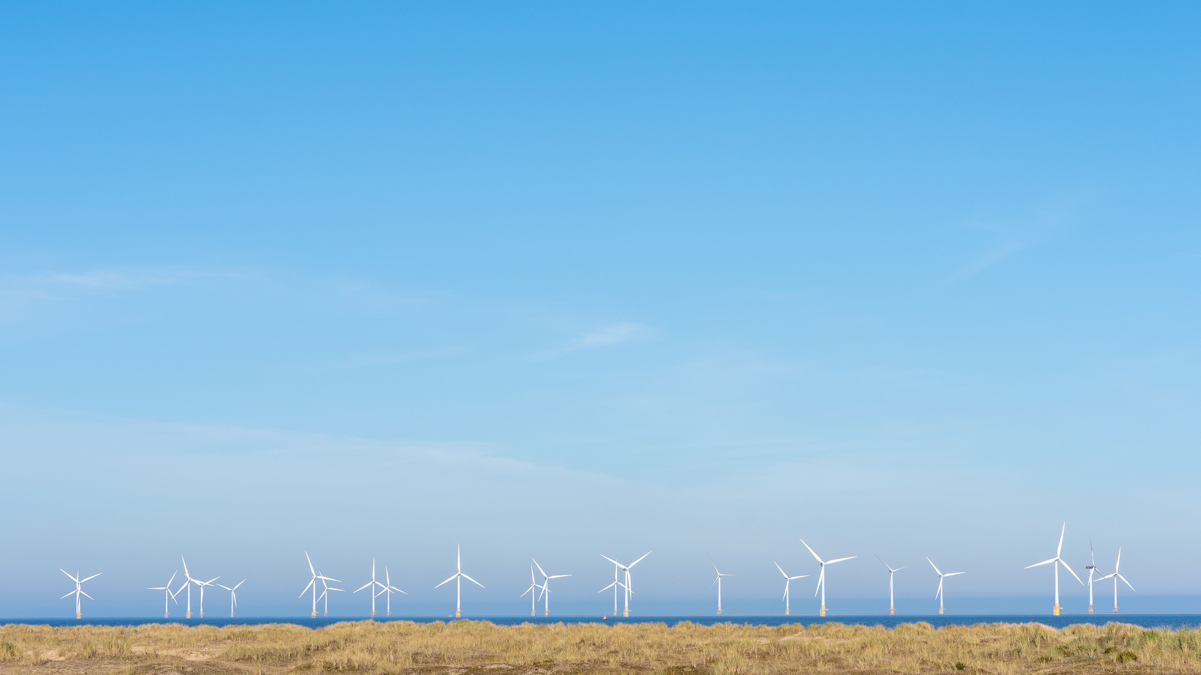 Offshore wind turbines on the horizon beyond sandy dunes under a clear blue sky