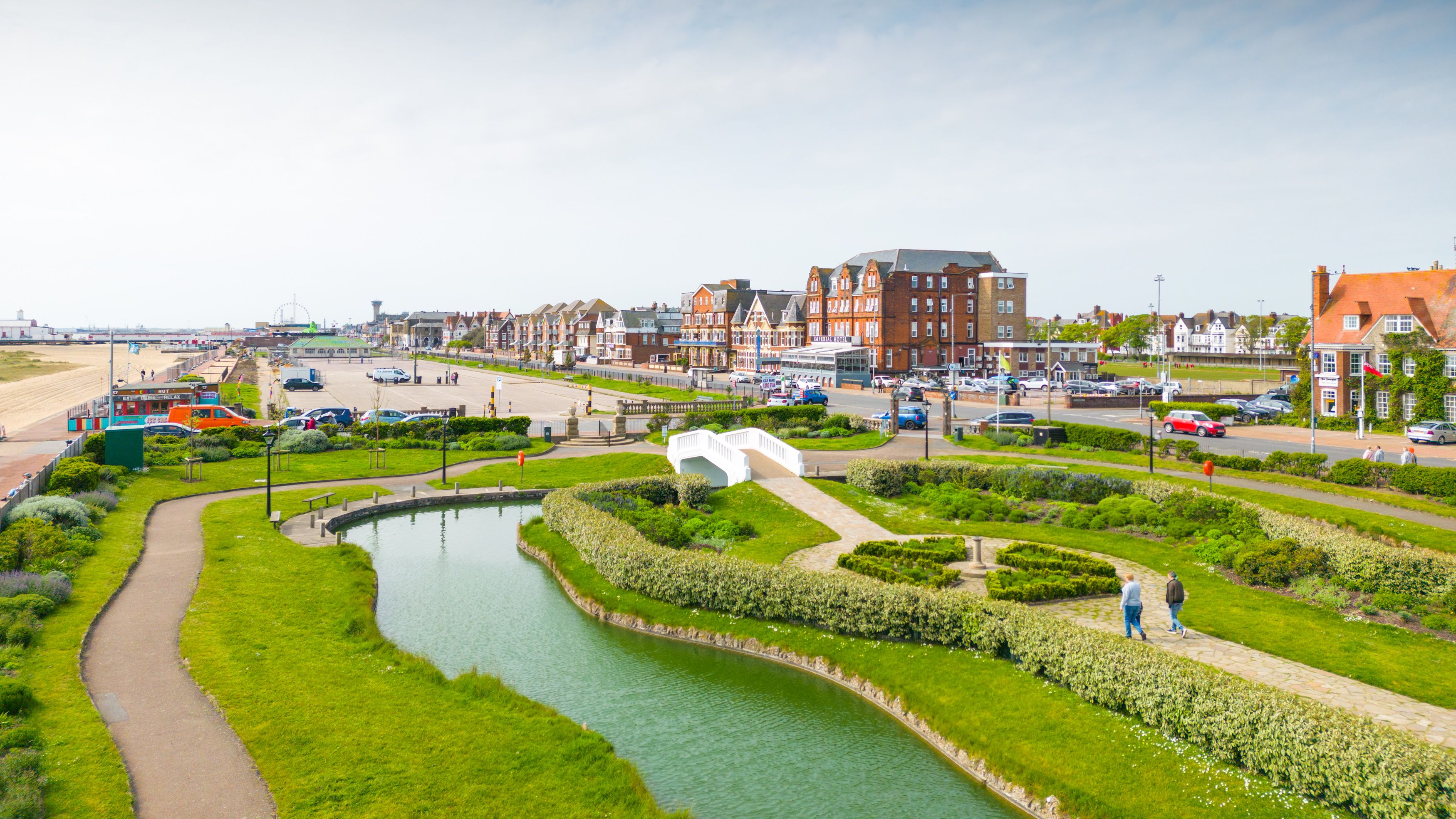 Seafront gardens with a small pond and white footbridge beside a coastal road and buildings