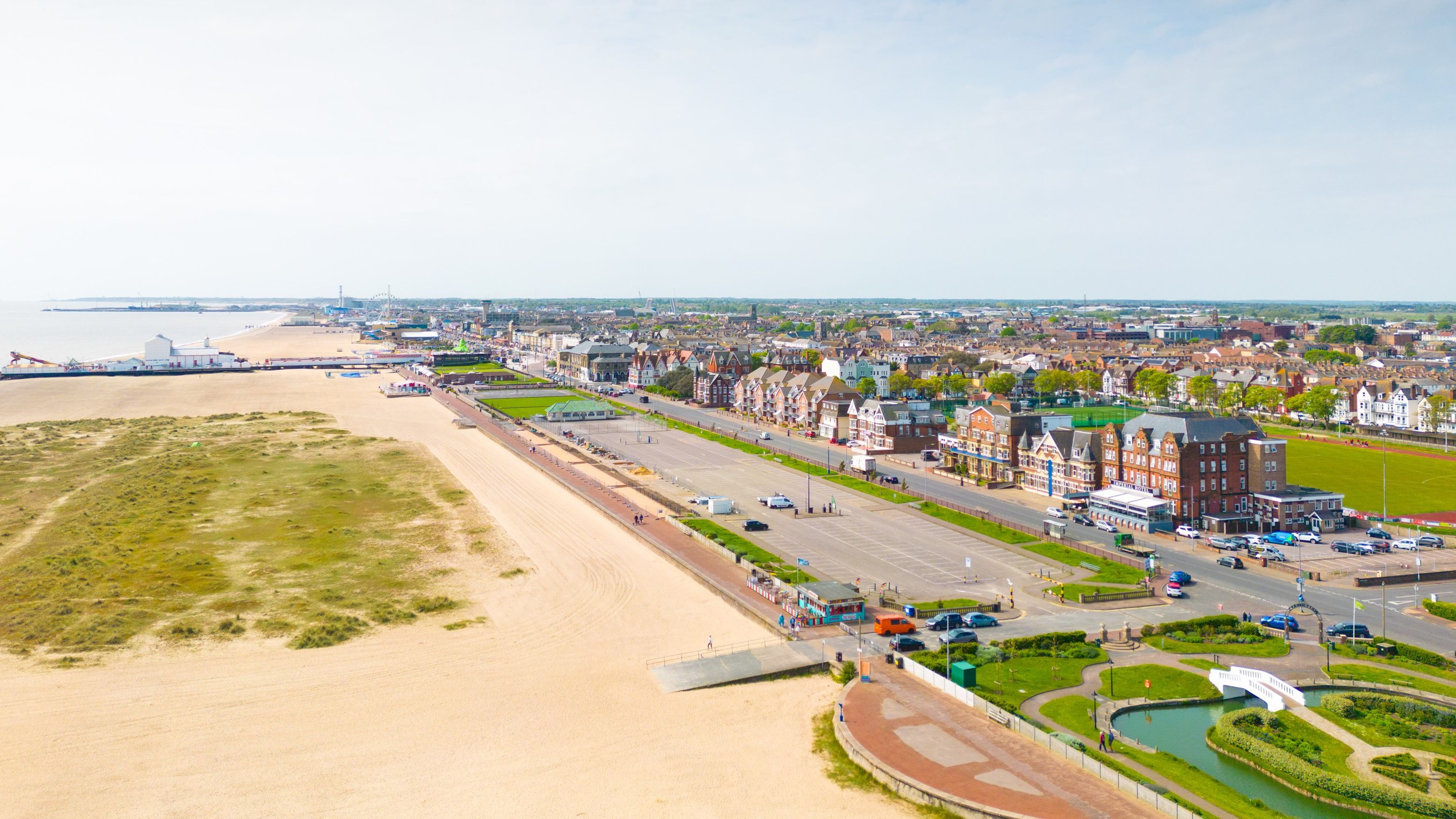 Aerial view of a seaside town with a long sandy beach, promenade, and rows of buildings under a clear sky.