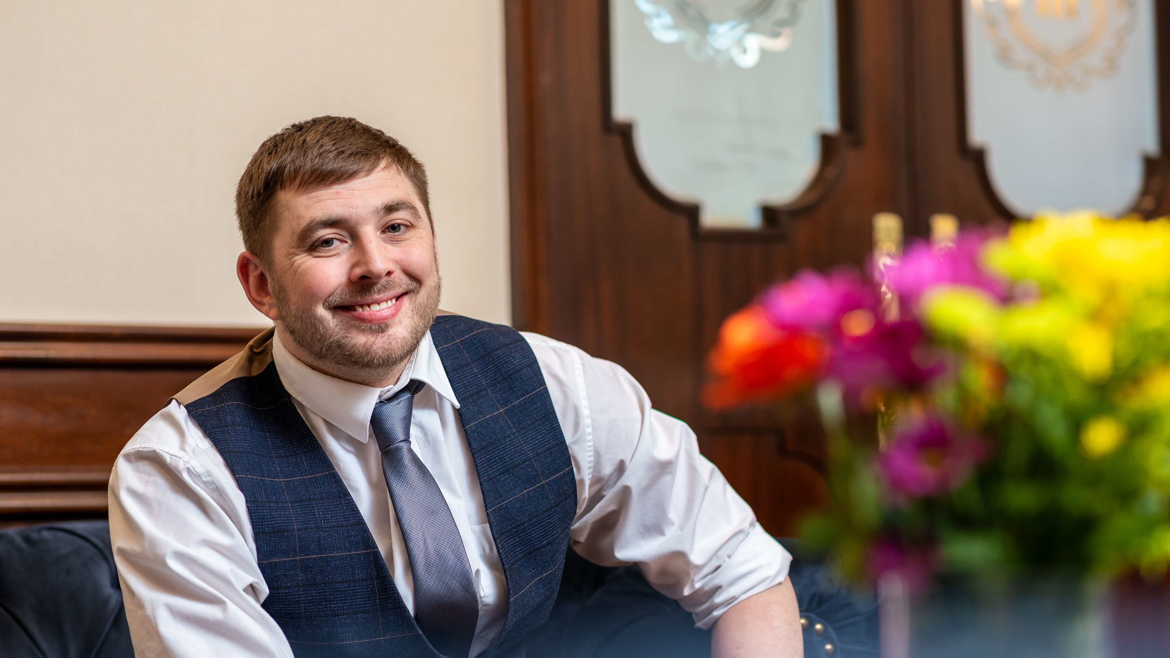 Smiling man in a waistcoat and tie seated in a hotel lounge with colourful flowers in the foreground