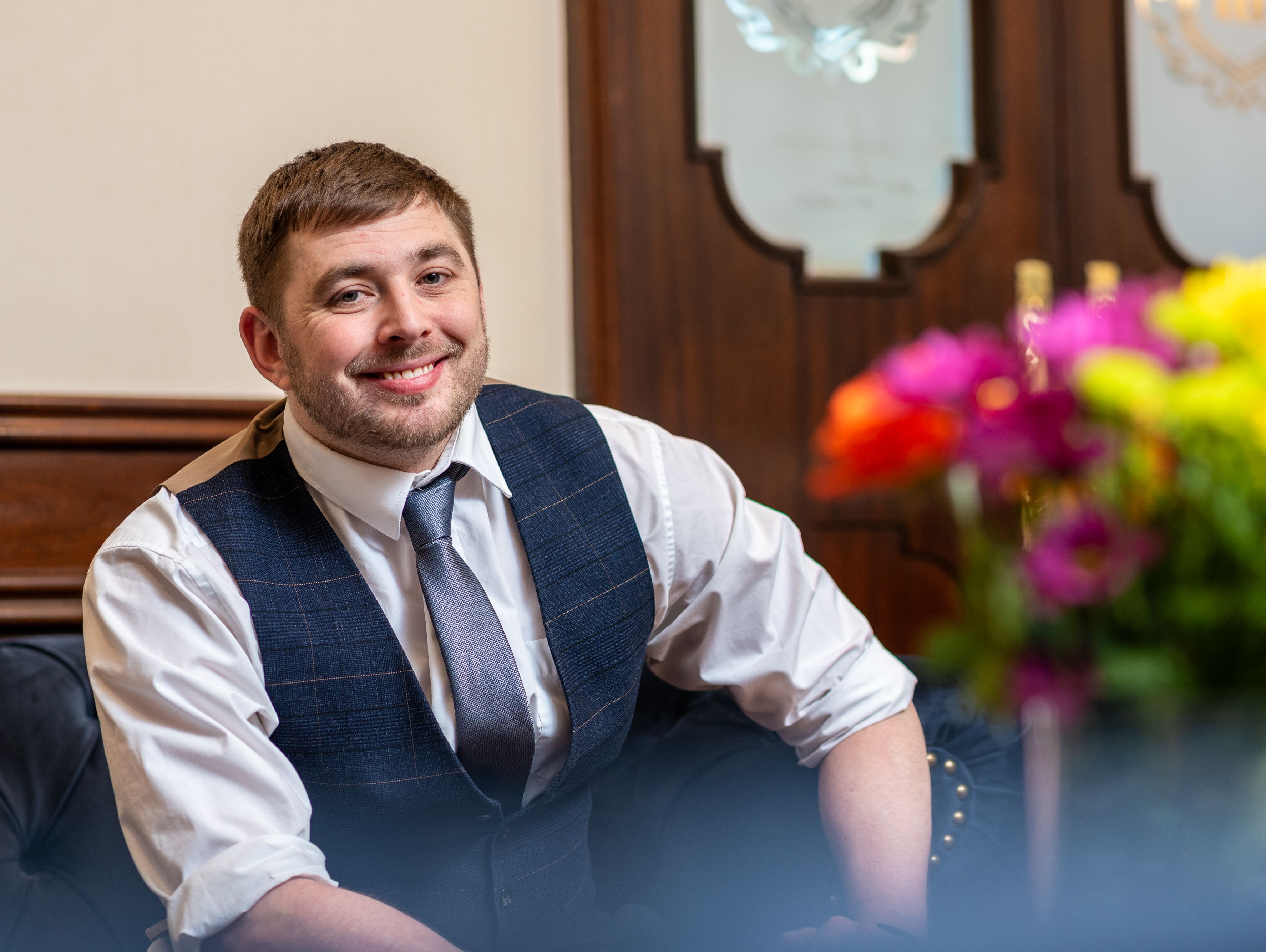 Smiling man in a waistcoat and tie seated in a hotel lounge with colourful flowers in the foreground