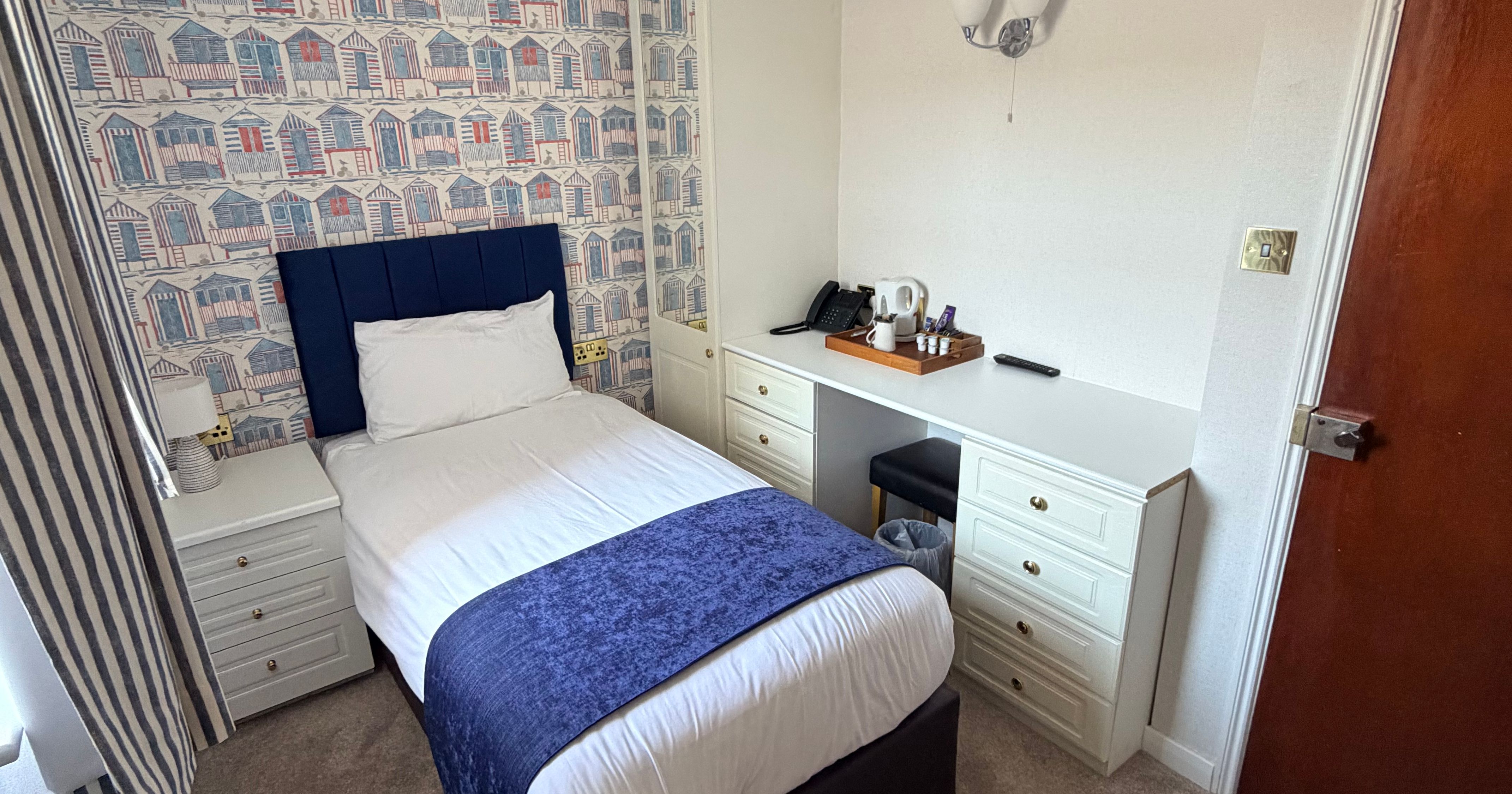 Single bedroom with patterned feature wallpaper, blue headboard, desk and tea tray