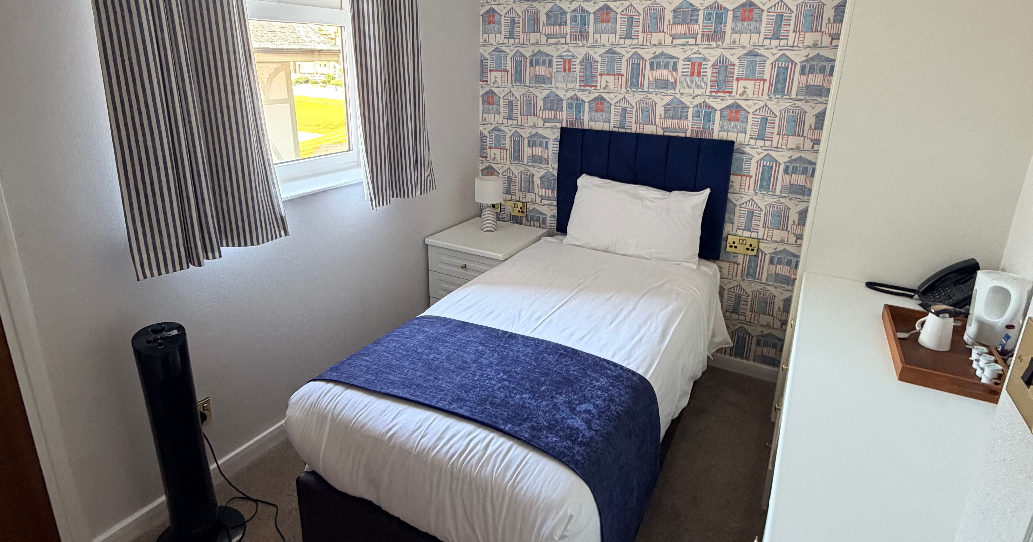 Compact single bedroom with navy headboard, striped curtains and seaside hut wallpaper