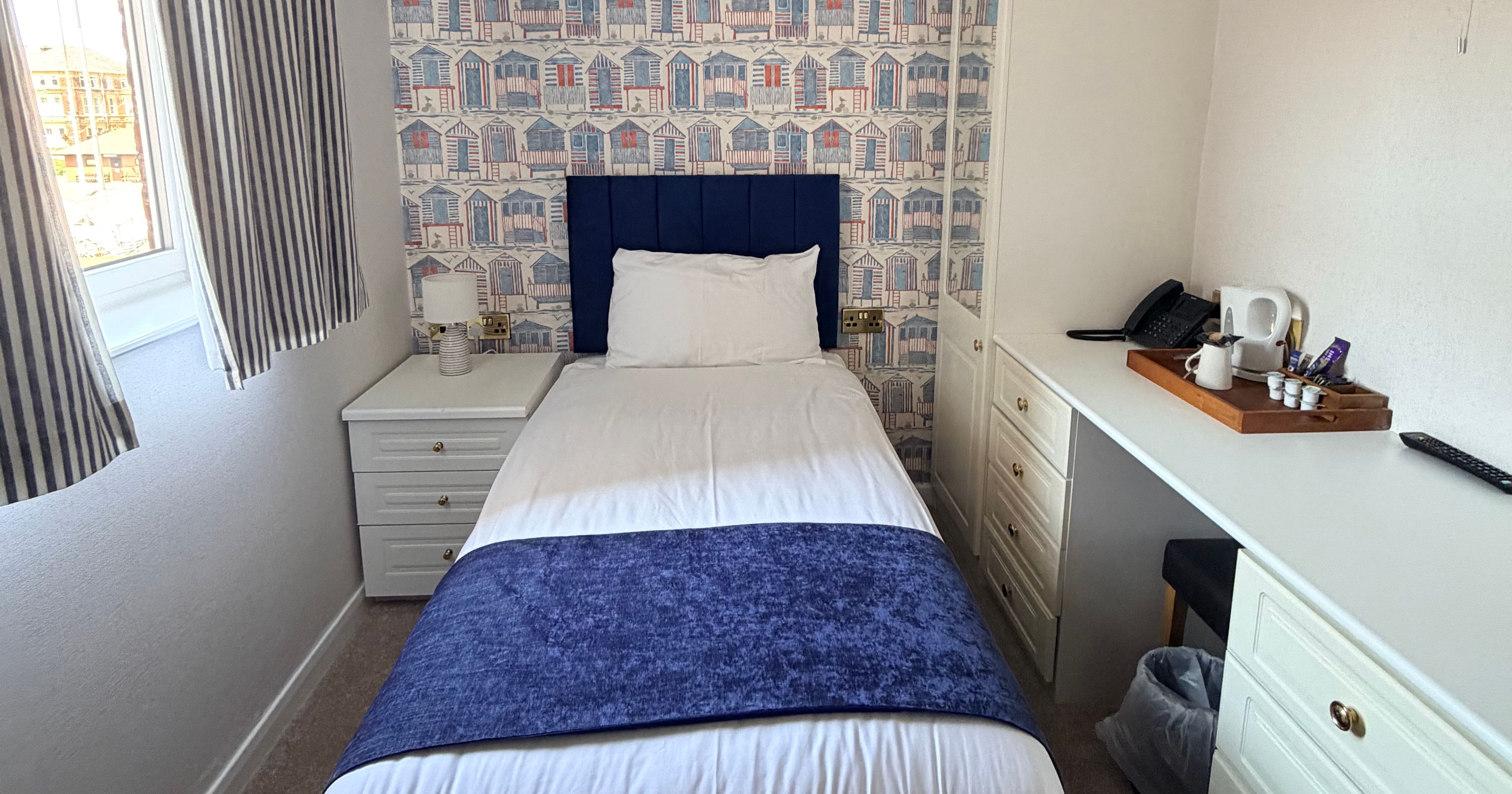 Single bed in a bright guest room with blue headboard, beach hut wallpaper and striped curtains