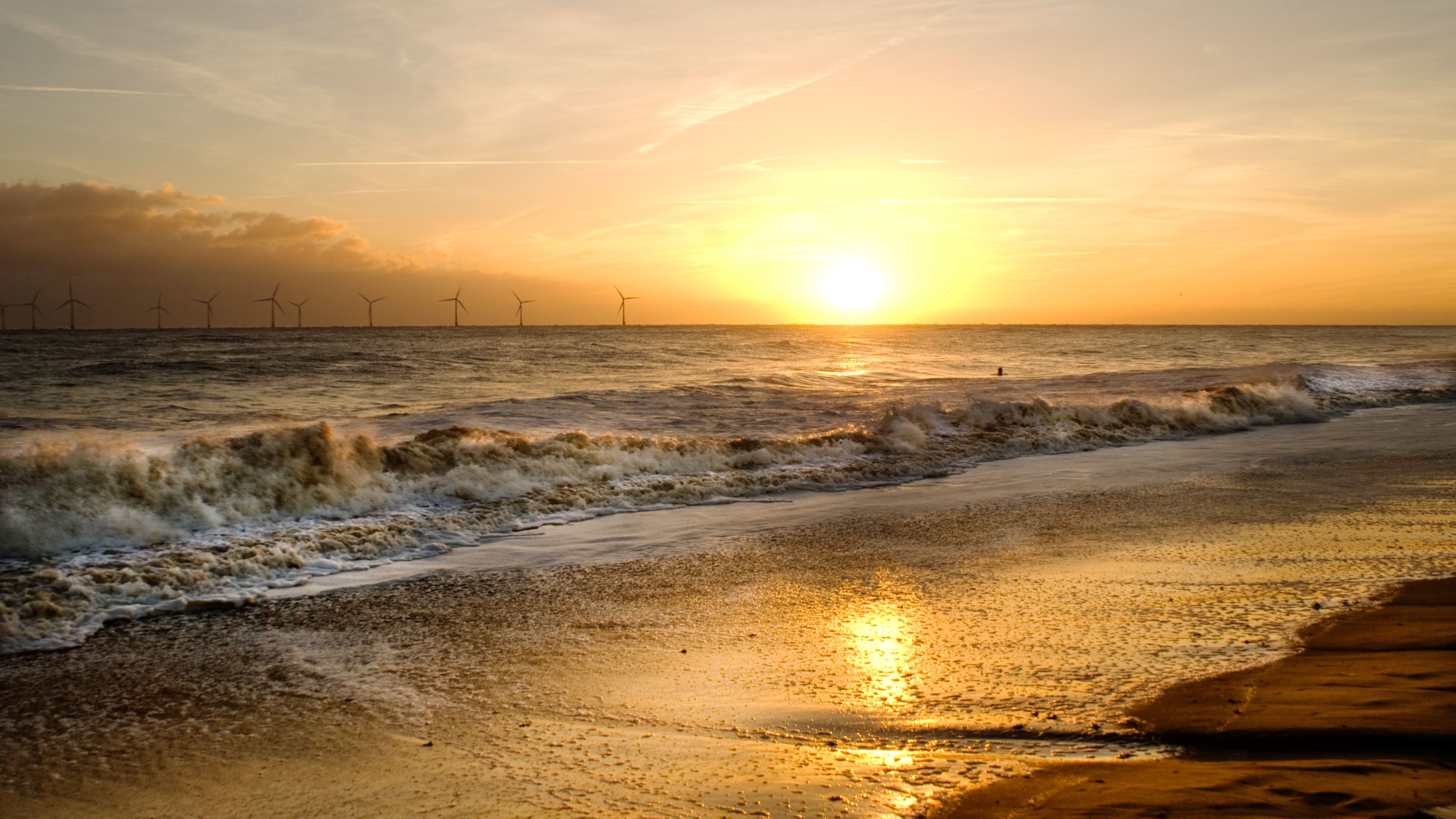 Golden sunset over a sandy beach with waves rolling in and wind turbines on the horizon