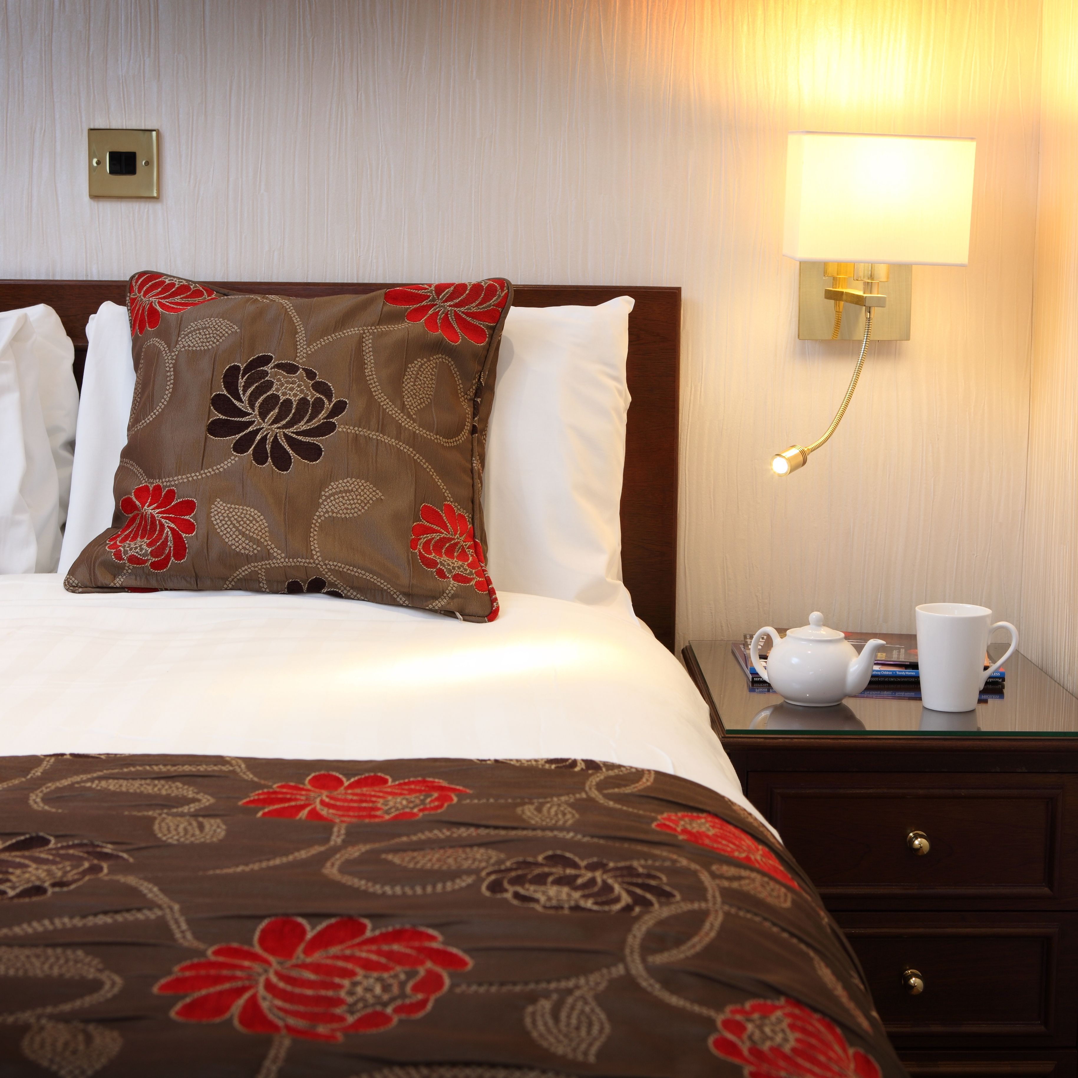 Hotel bed with floral cushions and bedside lamp next to a teapot and mug