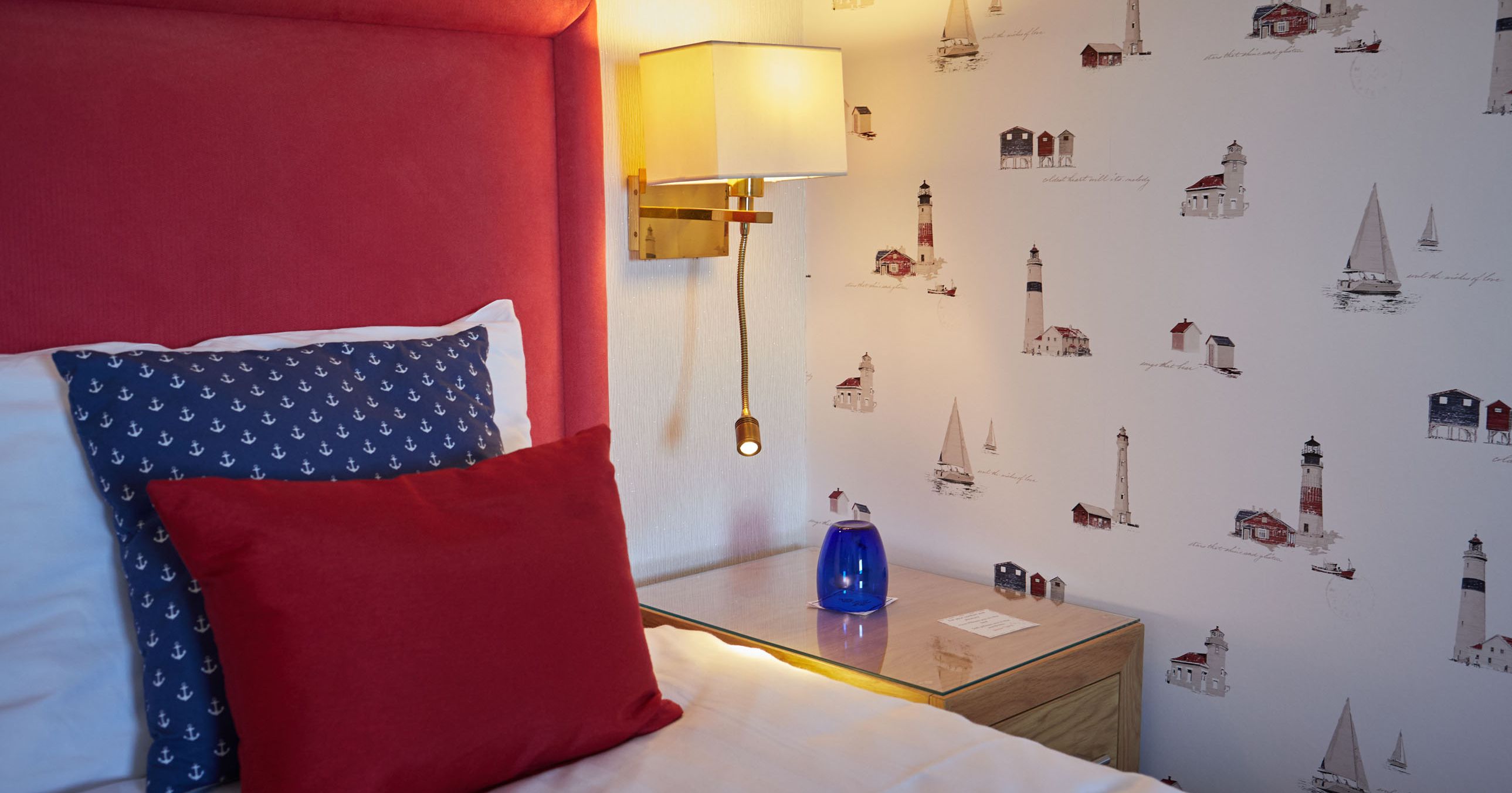 Cosy bedroom corner with red upholstered headboard, nautical cushions, bedside lamp and lighthouse-patterned wallpaper