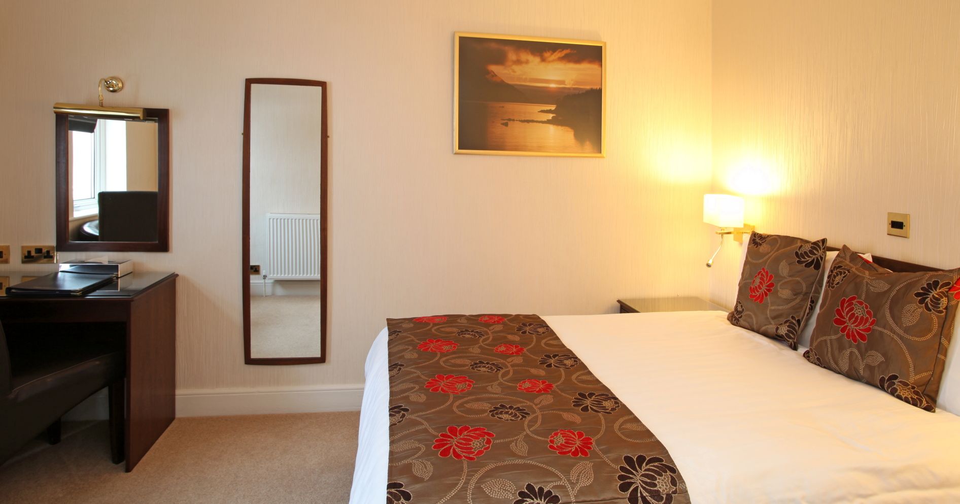 Cosy hotel bedroom with double bed, floral cushions, bedside lamp, mirrors and a framed sunset picture