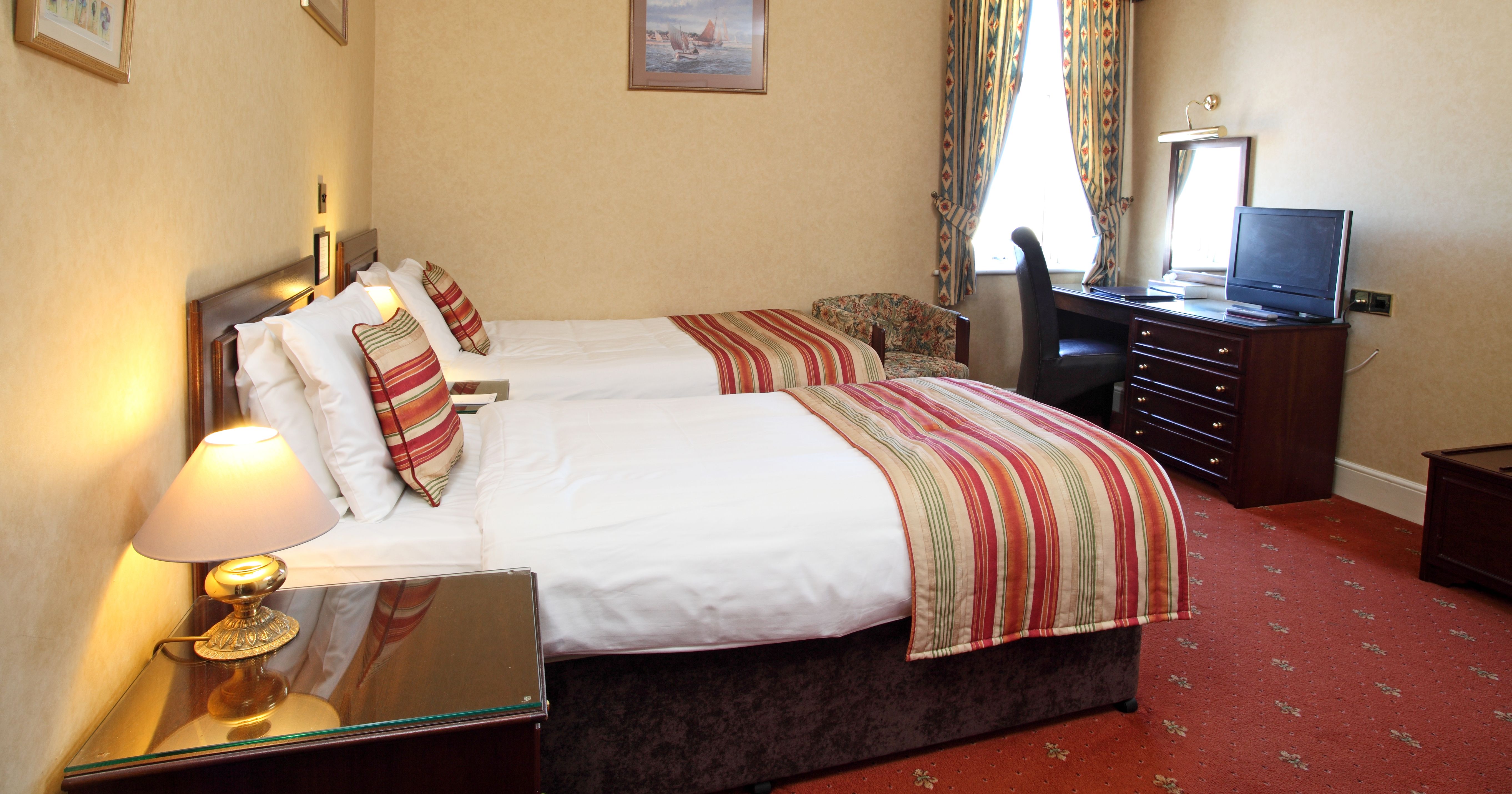 Hotel twin room with two single beds, striped bed runners, bedside lamp, and desk by a window with patterned curtains