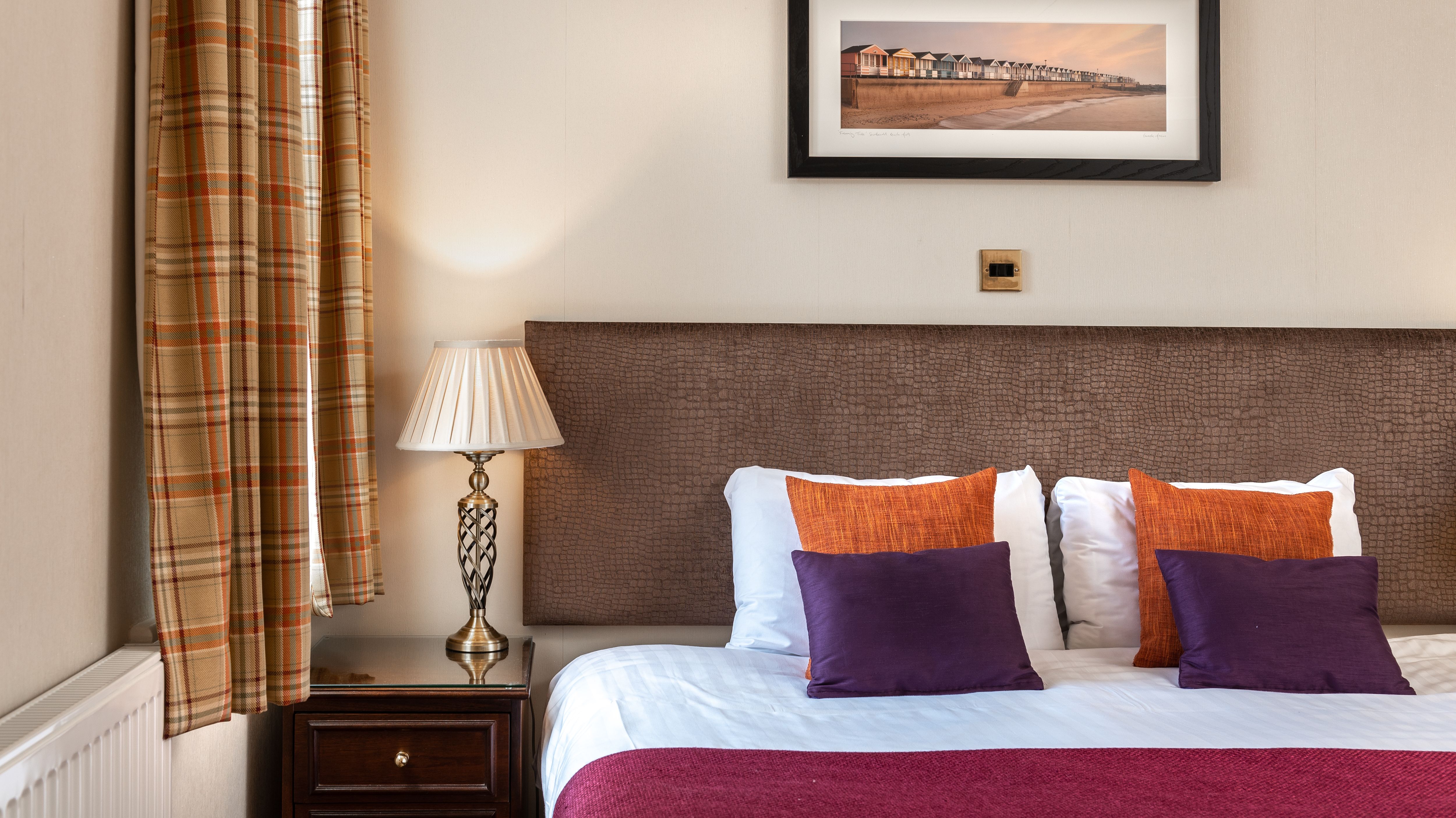 Cosy bedroom with double bed, purple and orange cushions, bedside lamp, and plaid curtains
