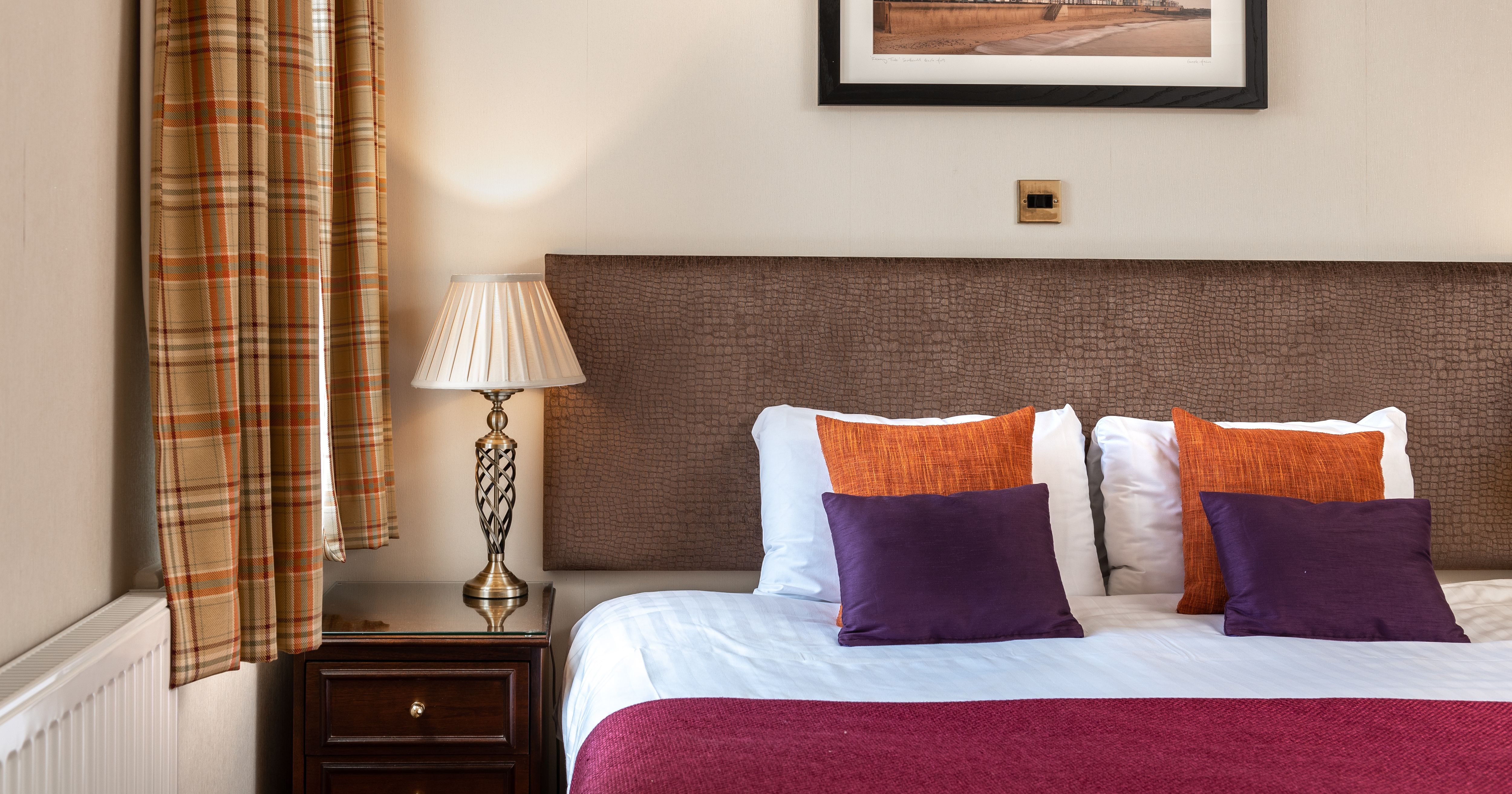 Cosy bedroom with double bed, purple and orange cushions, bedside lamp, and plaid curtains