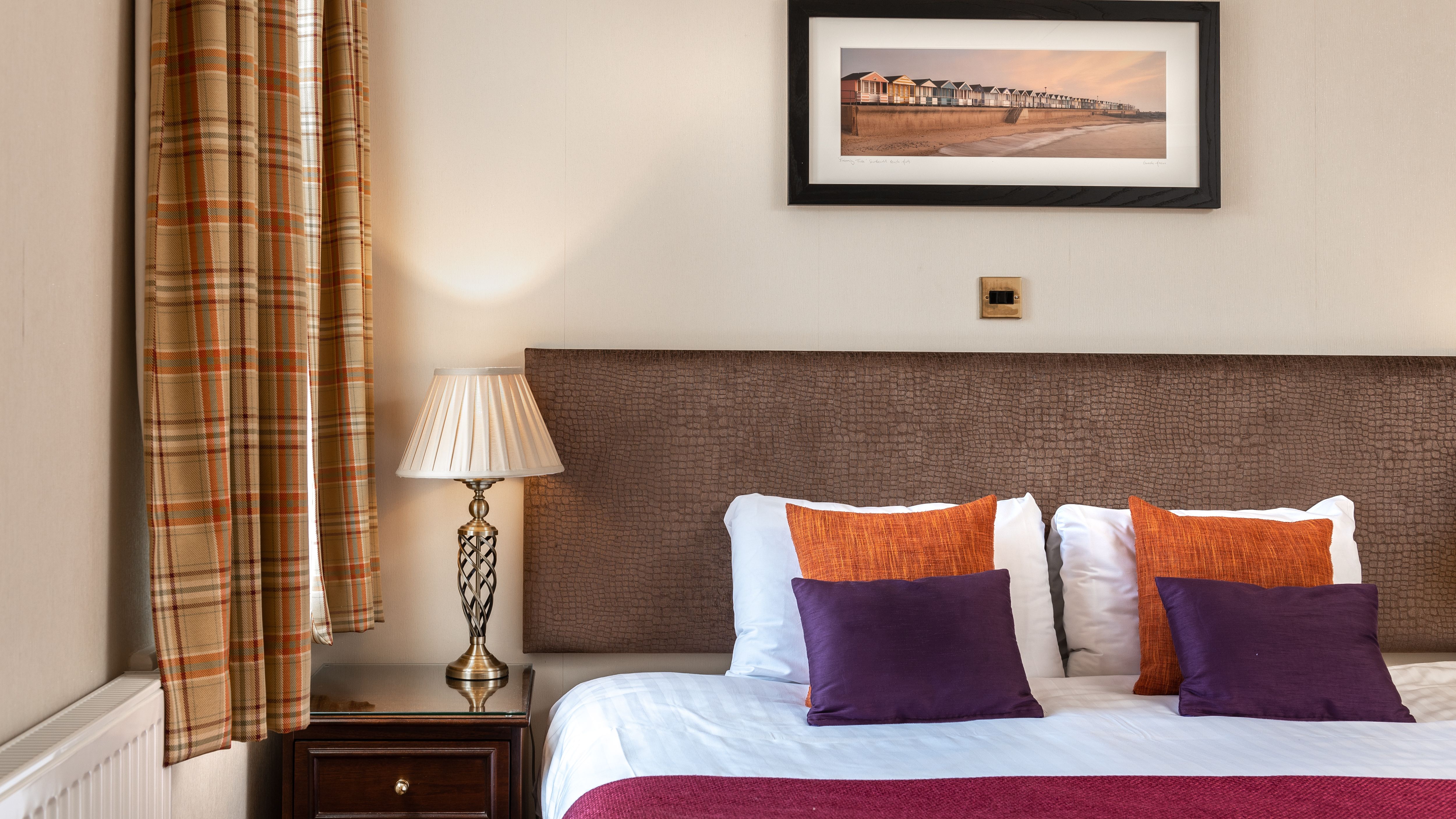 Cosy bedroom with double bed, purple and orange cushions, bedside lamp, and plaid curtains