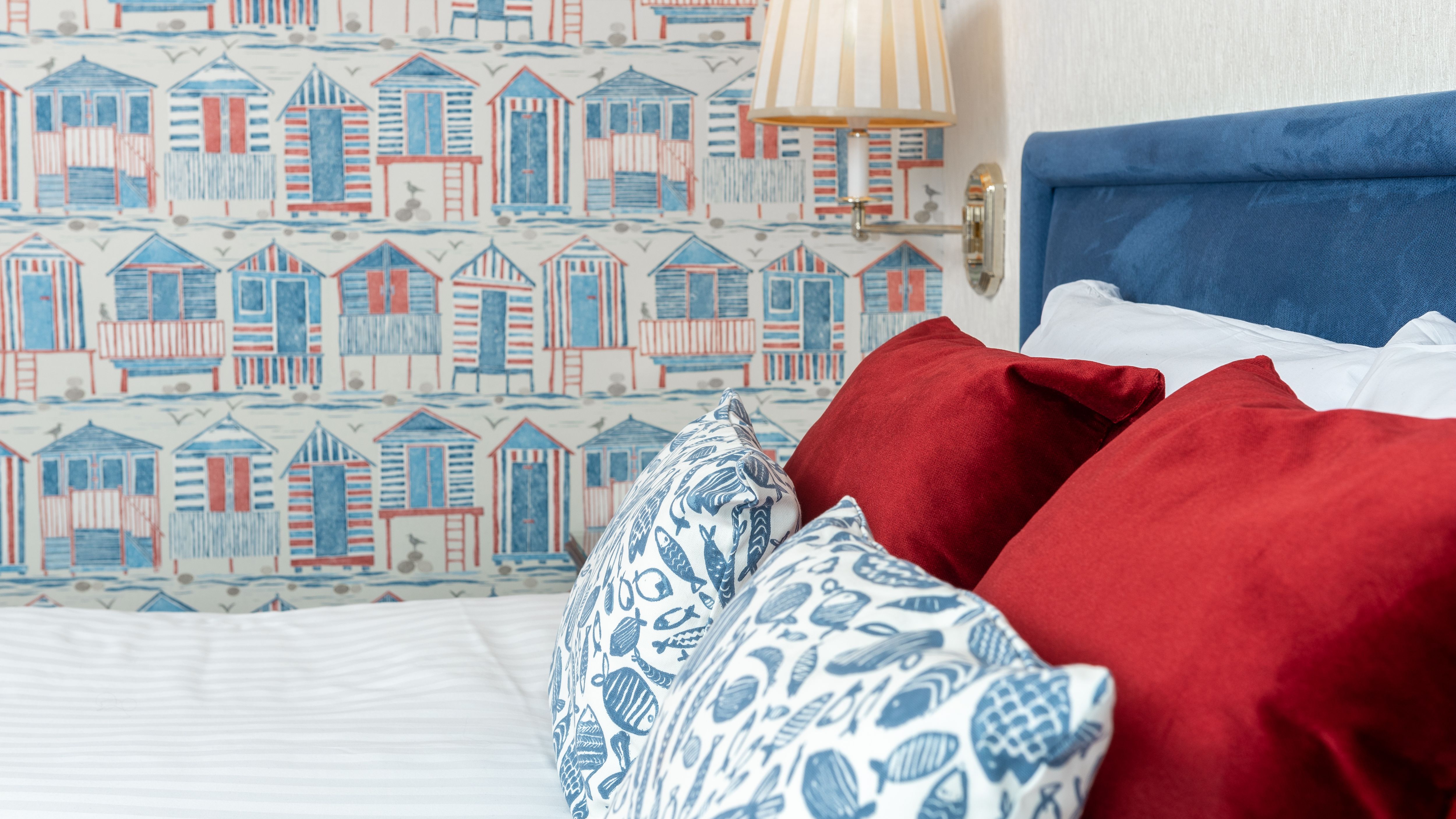 Bed with red and patterned cushions beside a blue headboard, a striped wall lamp and seaside hut wallpaper.