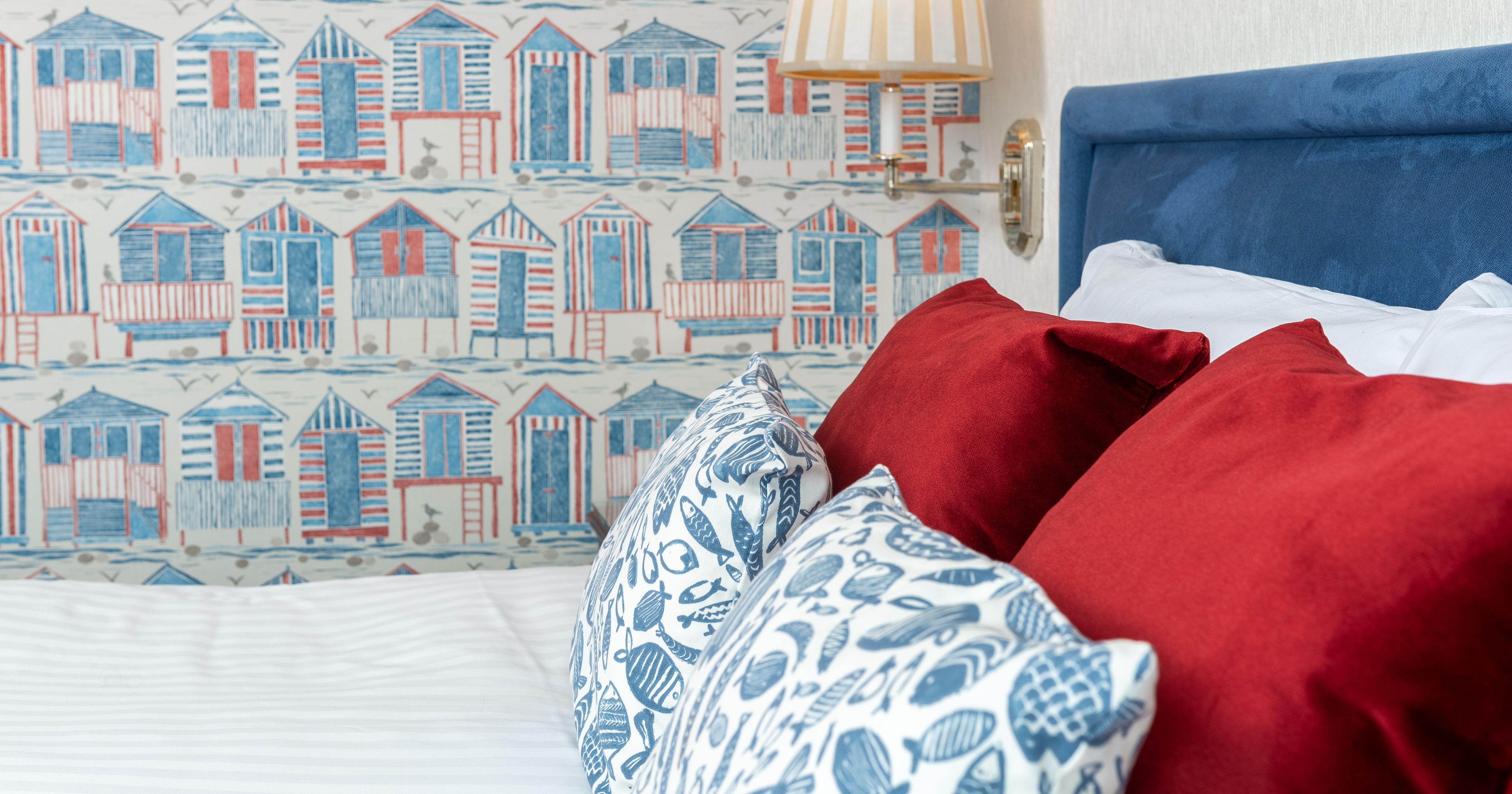 Bed with red and patterned cushions beside a blue headboard, a striped wall lamp and seaside hut wallpaper.