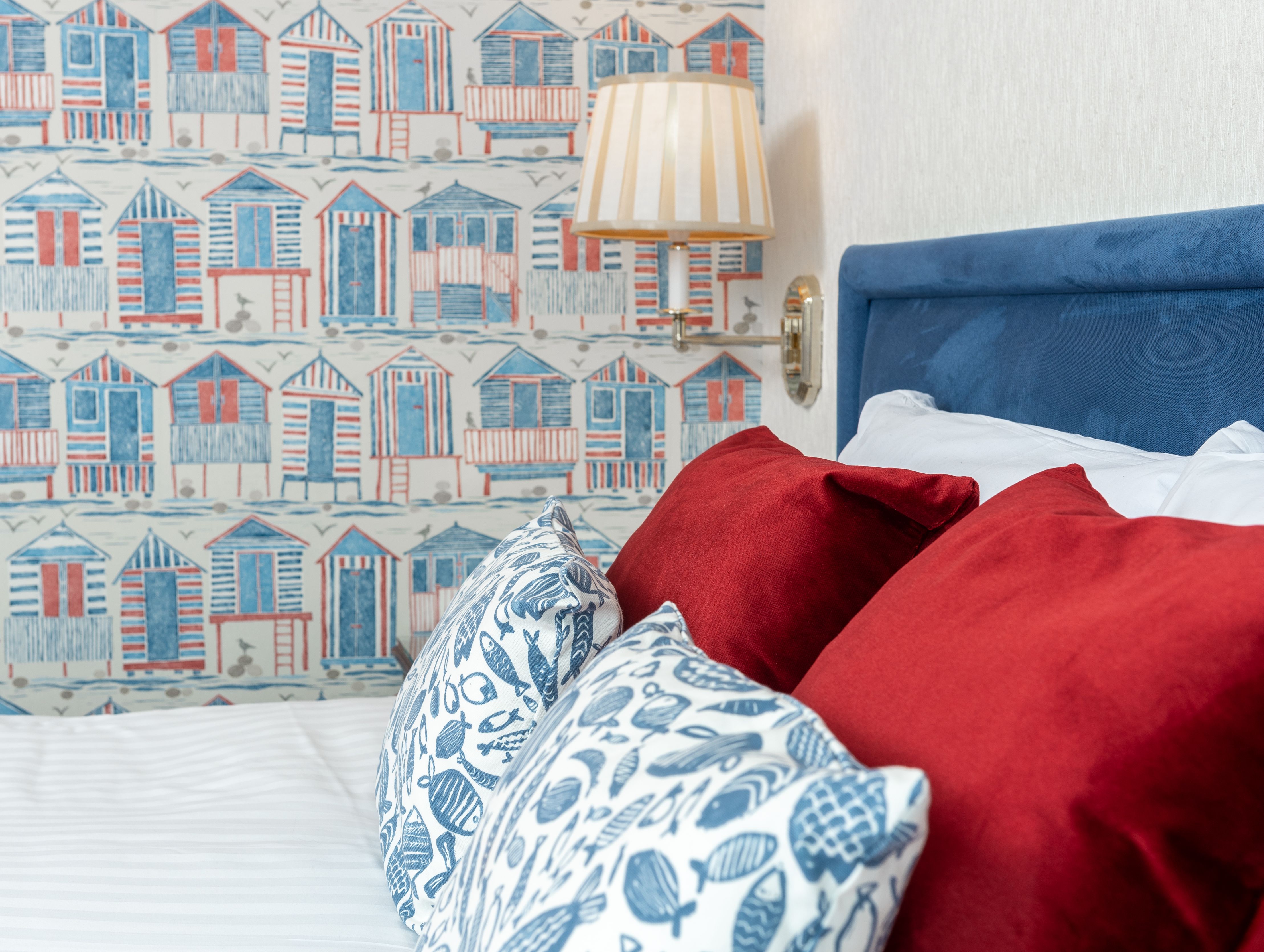 Bed with red and patterned cushions beside a blue headboard, a striped wall lamp and seaside hut wallpaper.