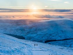 Two hikers walk a snowy path near Pont-ar-Daf in Bannau Brycheiniog, with rolling white hills and a glowing sunset casting warm tones across the frozen landscape.