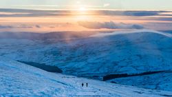 Two hikers walk a snowy path near Pont-ar-Daf in Bannau Brycheiniog, with rolling white hills and a glowing sunset casting warm tones across the frozen landscape.