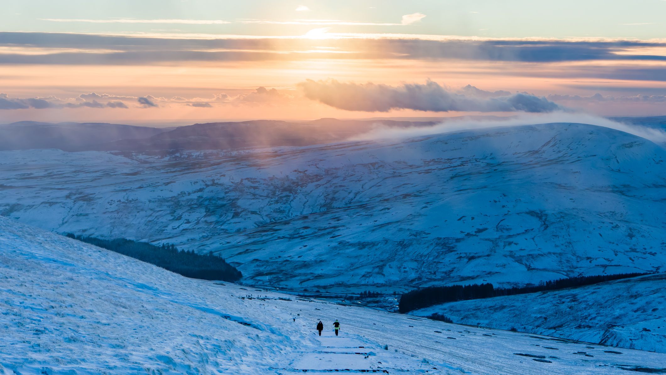 Two hikers walk a snowy path near Pont-ar-Daf in Bannau Brycheiniog, with rolling white hills and a glowing sunset casting warm tones across the frozen landscape.