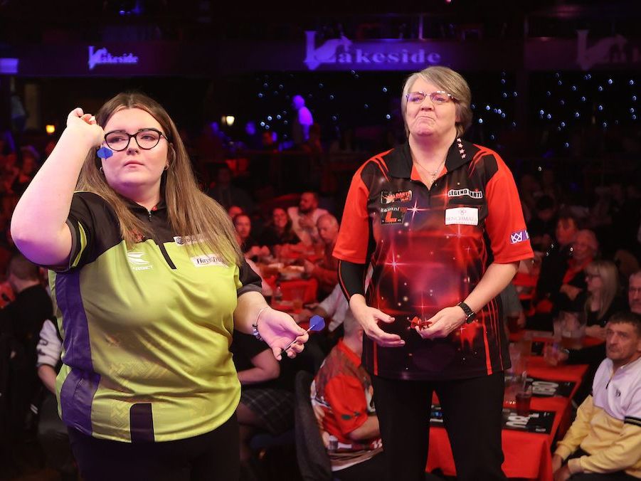 Two women compete in a darts match on stage in front of an audience.