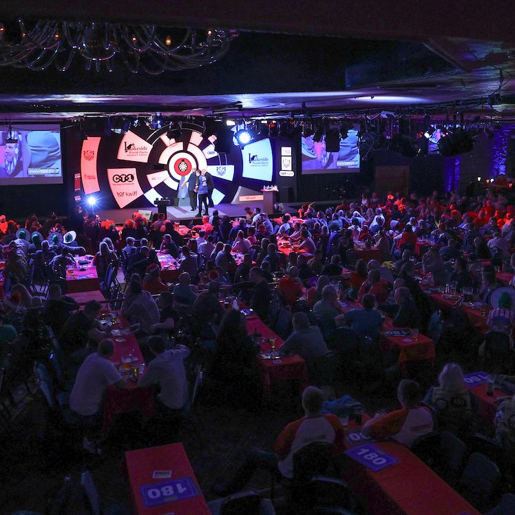 Large crowd watching a darts competition at a well-lit venue with a stage and dartboard in front.