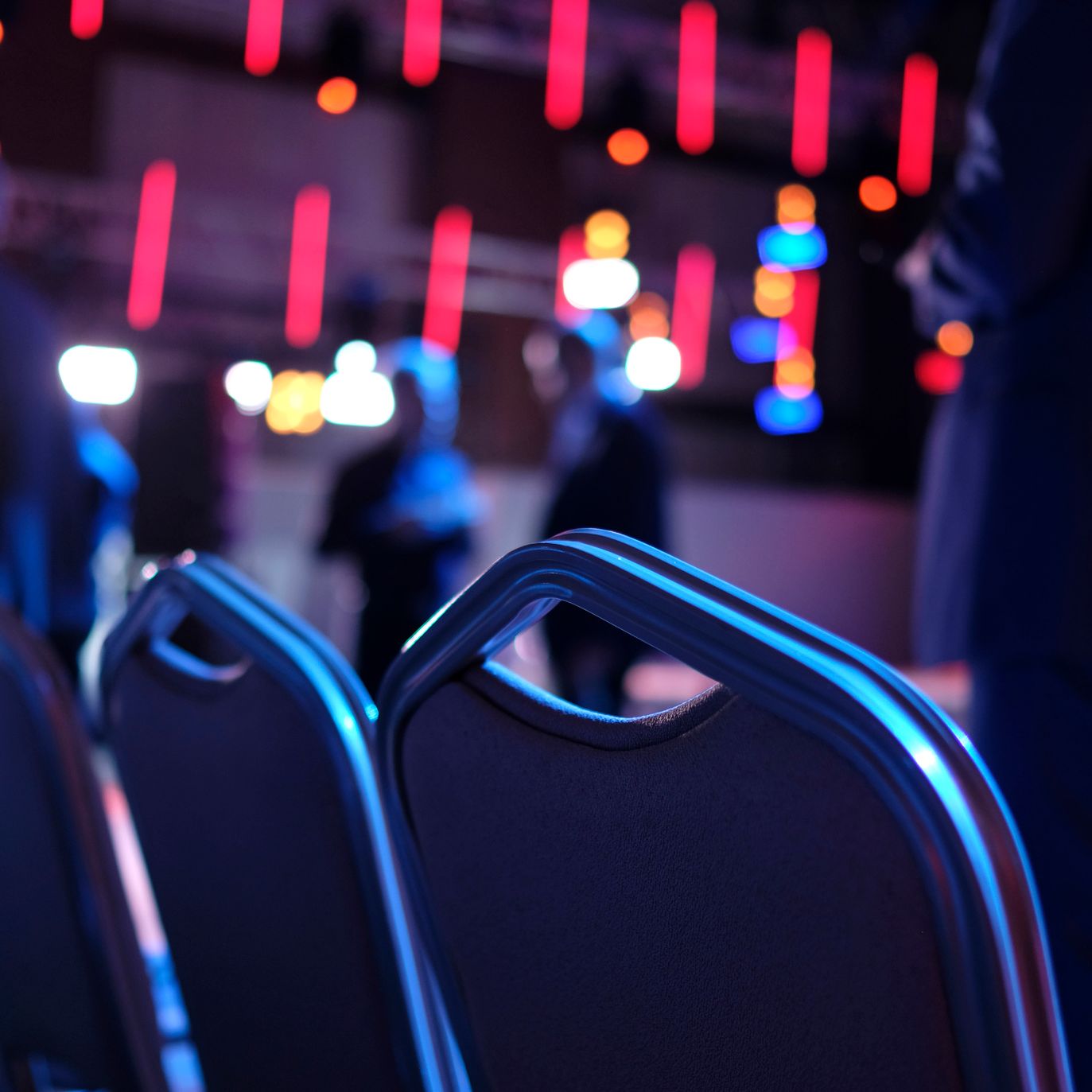 Rows of chairs in focus with people standing and colorful stage lights blurred in the background, suggesting an event or conference setting.