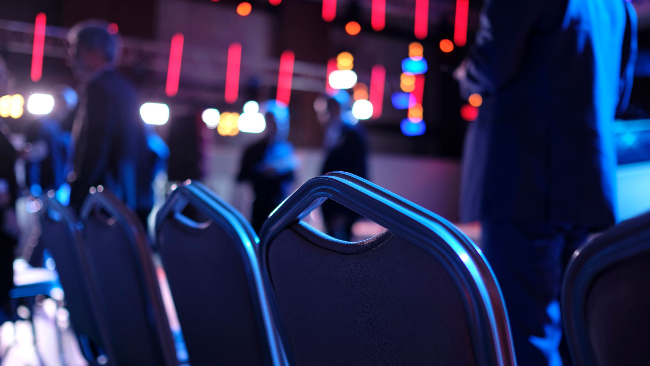 Rows of chairs in focus with people standing and colorful stage lights blurred in the background, suggesting an event or conference setting.