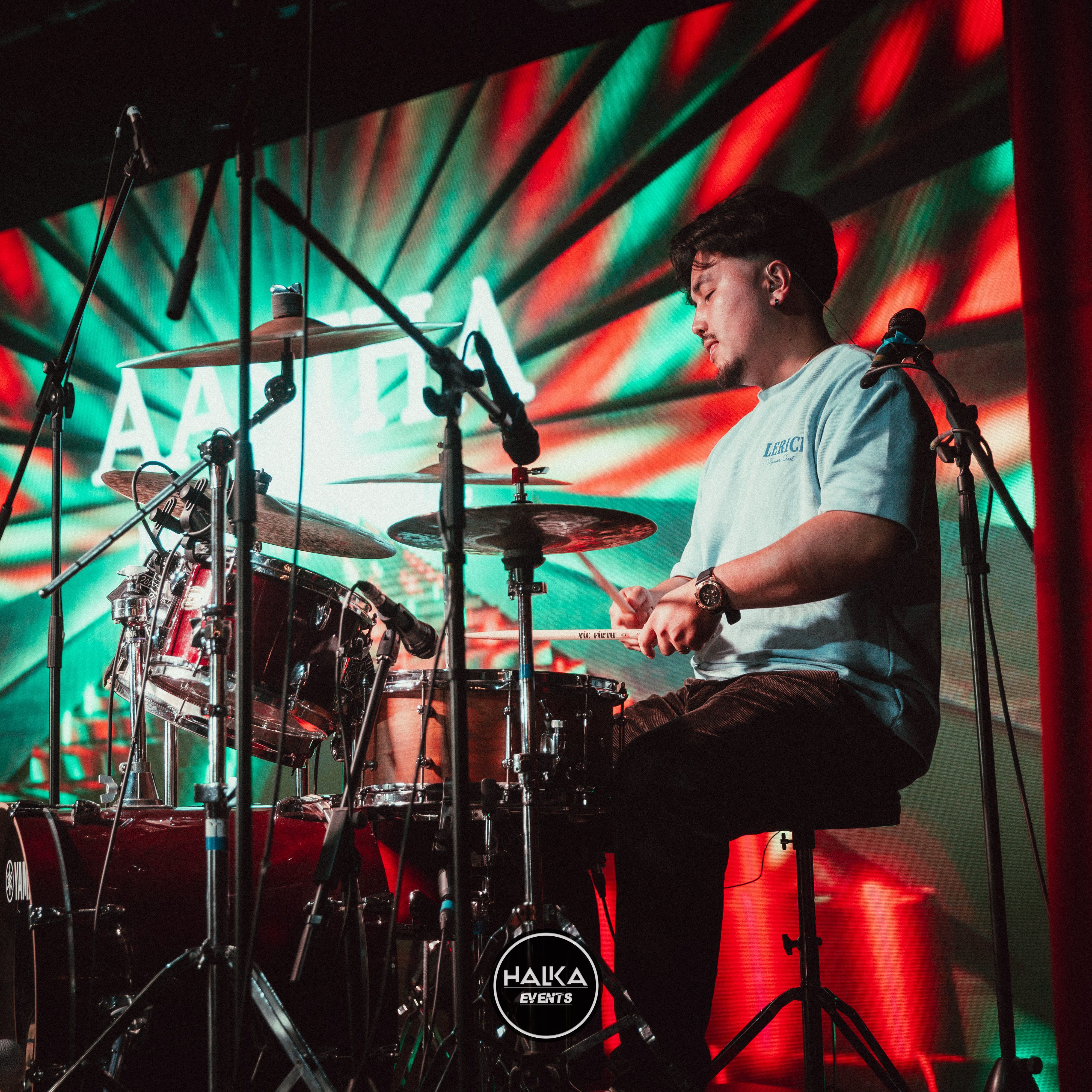 Drummer playing a drum kit on stage with red and green lights and microphones around the set