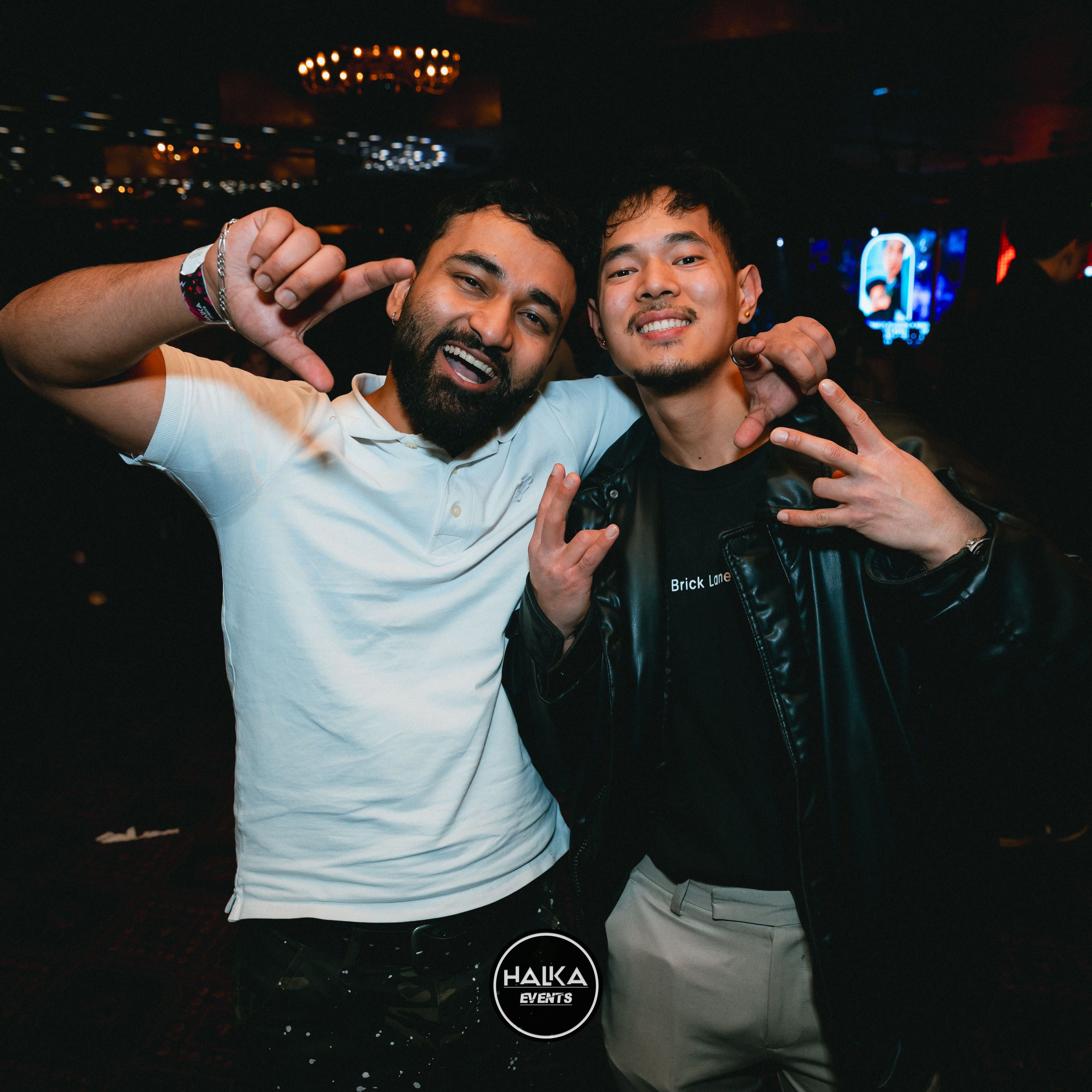 Two friends posing and smiling at a nightclub event, making hand signs under warm lights.